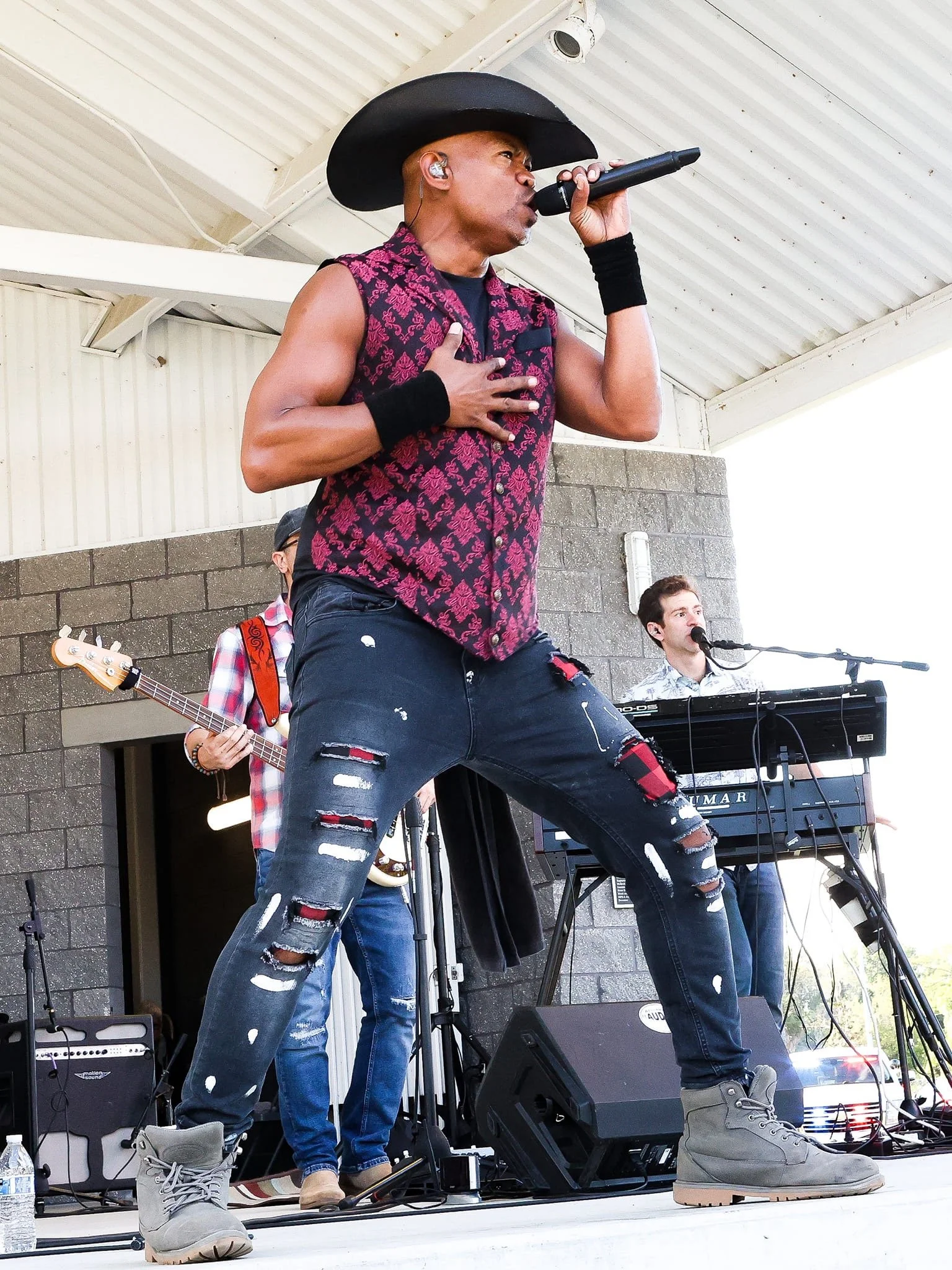 A singer wearing a cowboy hat and sleeveless patterned shirt performs on stage, gripping a microphone. The musician is accompanied by a band with a bassist, keyboardist, and visible equipment like speakers and a water bottle.
