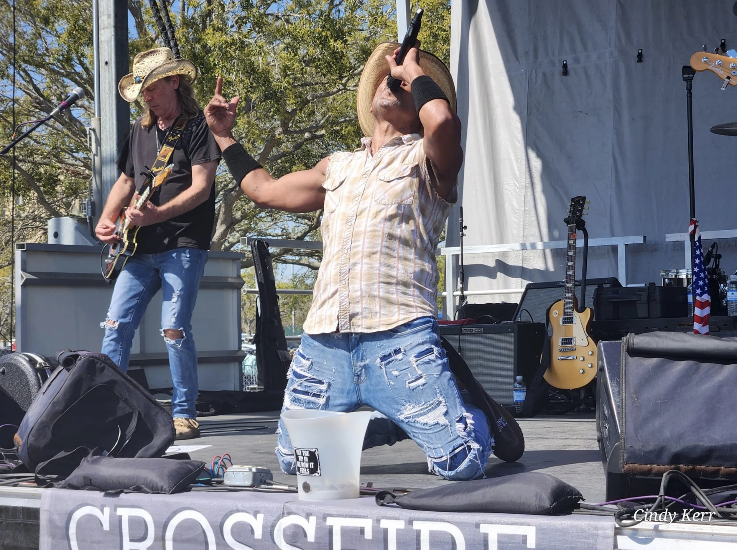 Musicians performing on an outdoor stage; one is playing electric guitar and the other singing into a microphone while kneeling. The stage is equipped with instruments and sound equipment. Both are wearing casual attire including jeans and hats.
