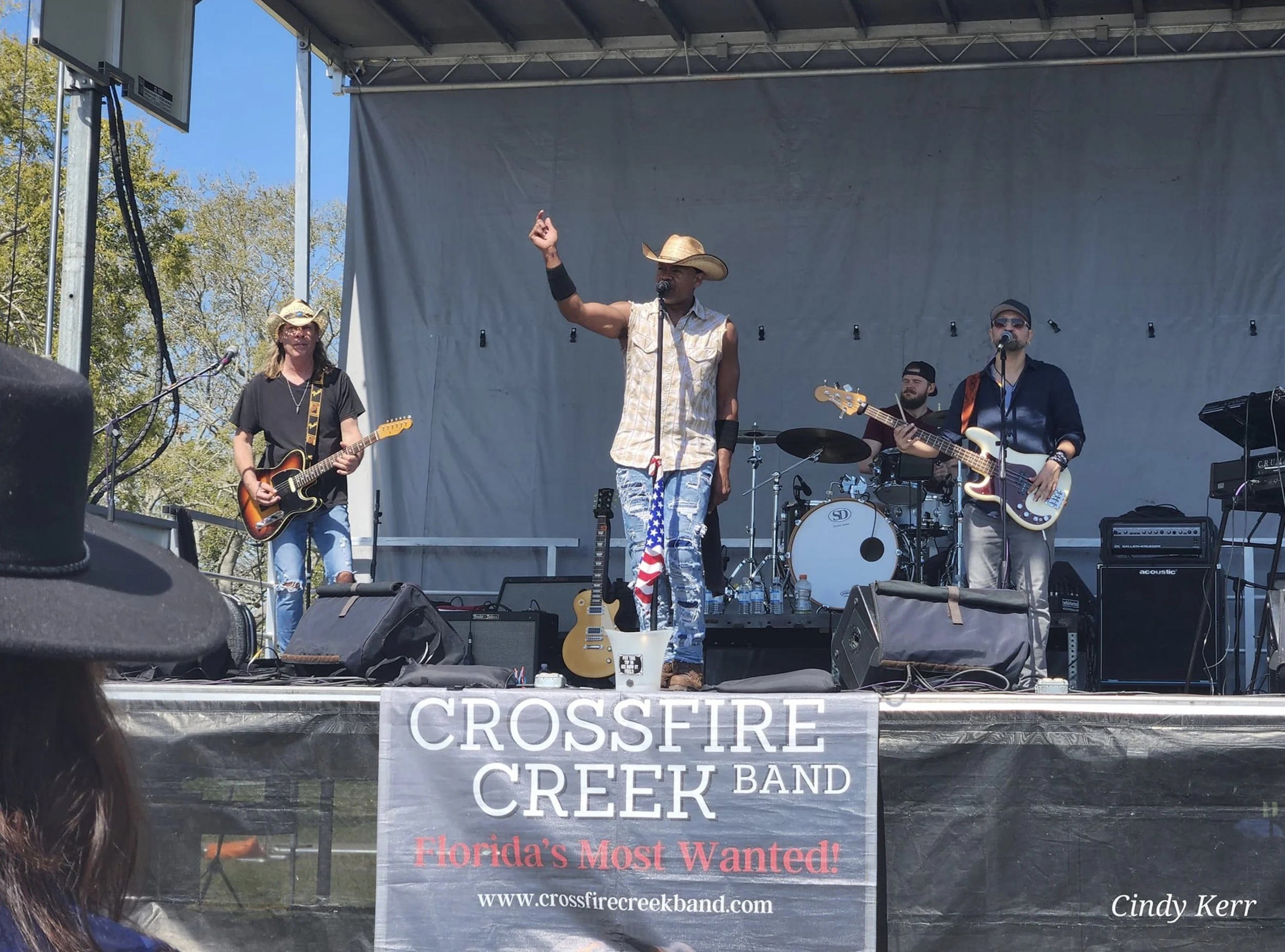Band performing on an outdoor stage with a "Crossfire Creek Band" sign and instruments, including guitars and drums.