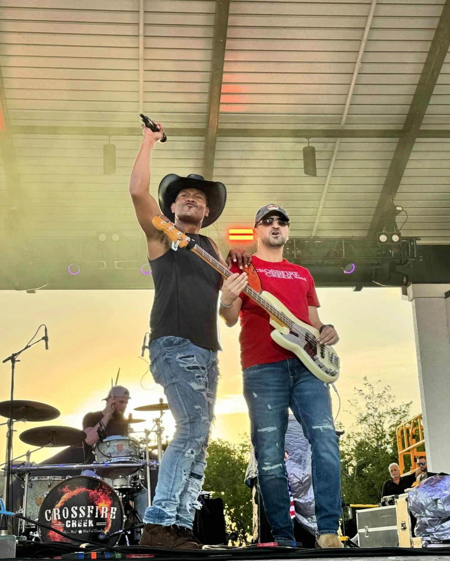 Three musicians performing on an outdoor stage. The one on the left is wearing a cowboy hat and holding a microphone and bass guitar. The center musician plays a bass guitar and wears a red shirt. The drummer is in the background. The drum kit has th
