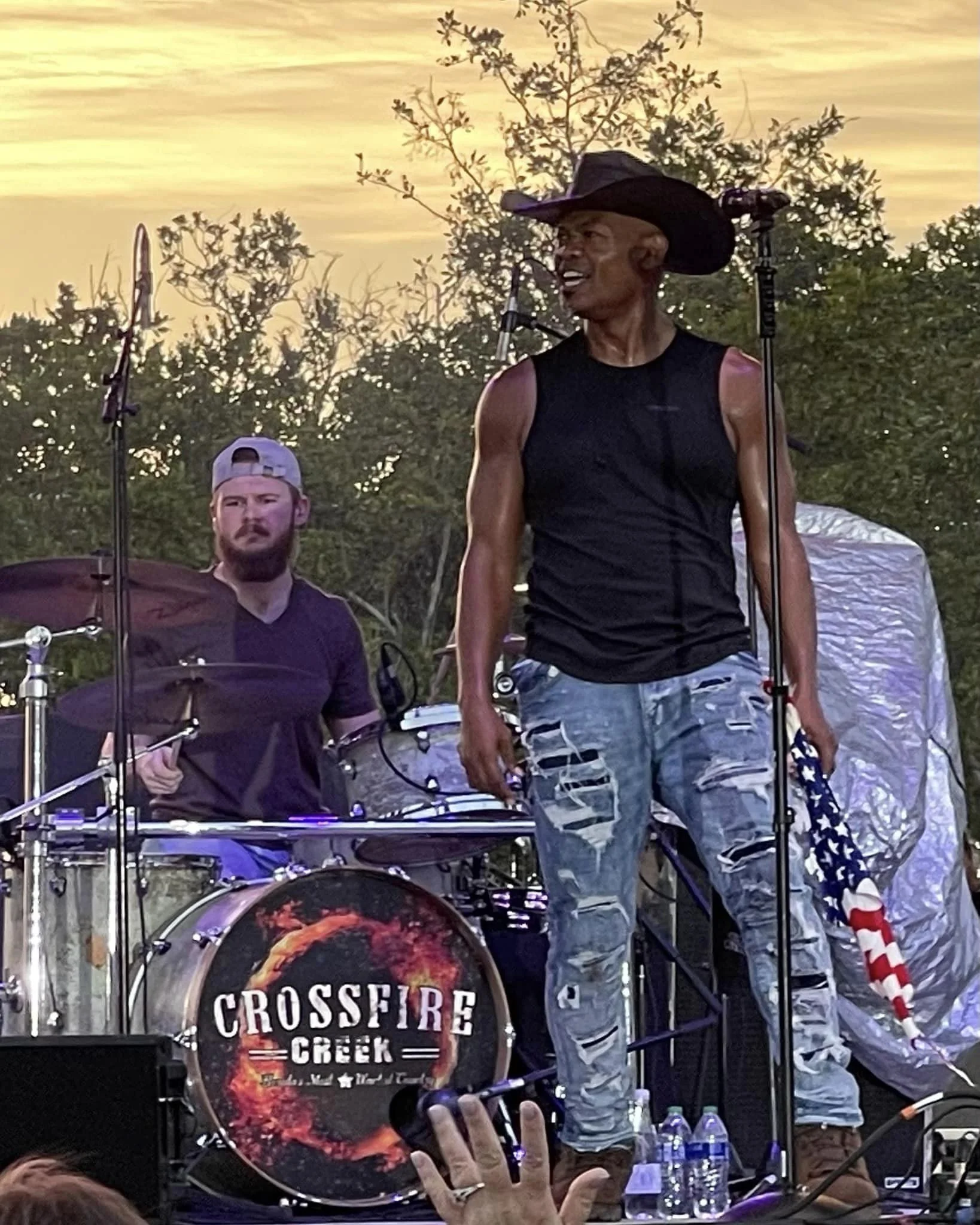 Musicians performing live, with a drummer and a vocalist wearing a cowboy hat and holding an American flag scarf, on an outdoor stage at sunset. Drum kit with 'Crossfire Creek' logo visible.