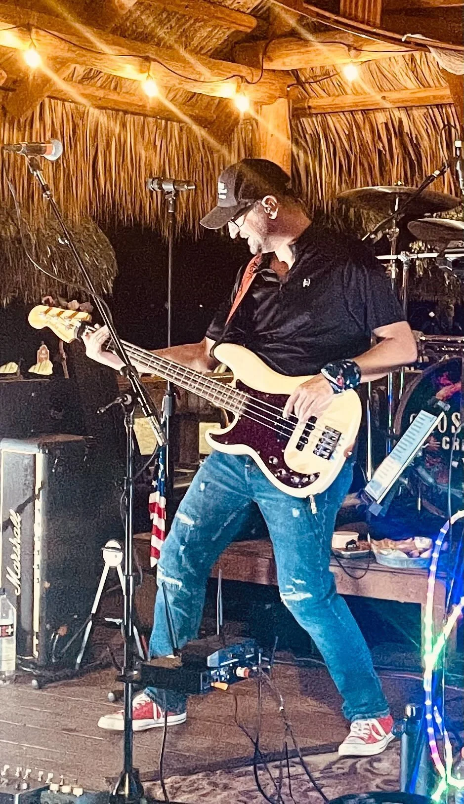 Musician playing guitar on stage with drum set and microphone, red shoes, outdoor setting with string lights.