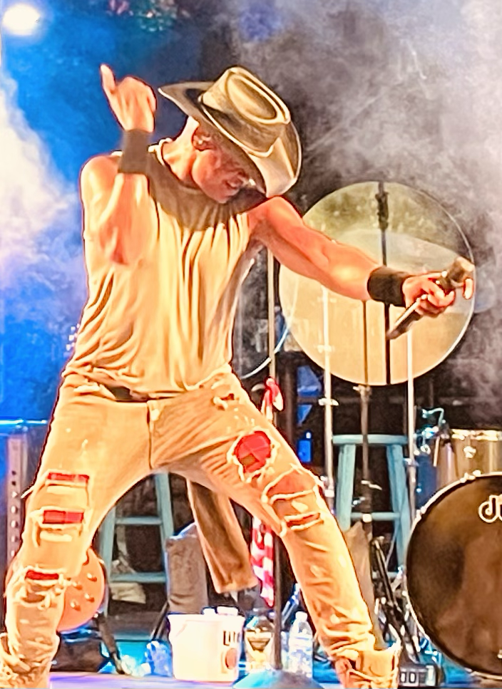 Performer with cowboy hat singing energetically on stage with microphone, wearing ripped jeans and a sleeveless shirt, vibrant stage lights in the background.
