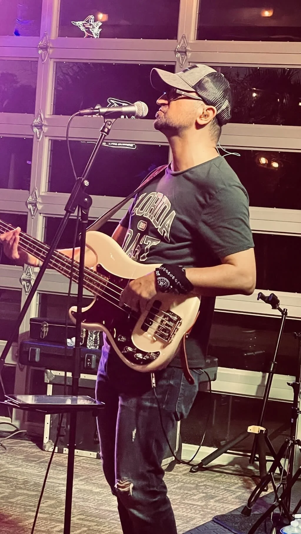Musician playing electric bass guitar and singing into microphone, wearing a cap and t-shirt.