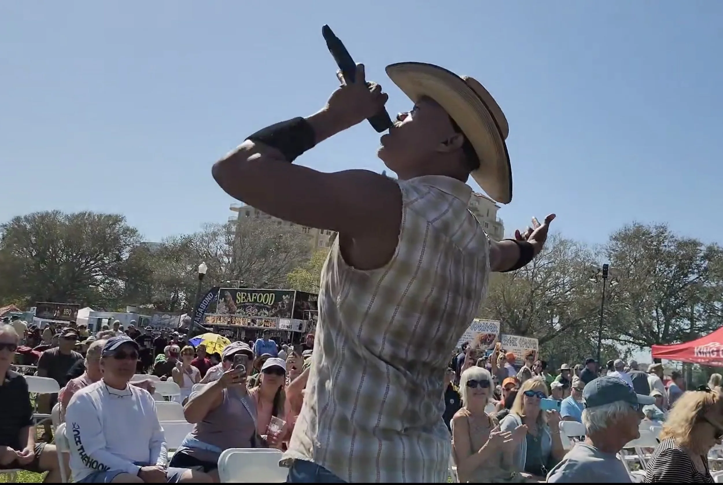 Person wearing a cowboy hat holding a microphone, performing in front of a seated outdoor audience, with food stalls in the background.