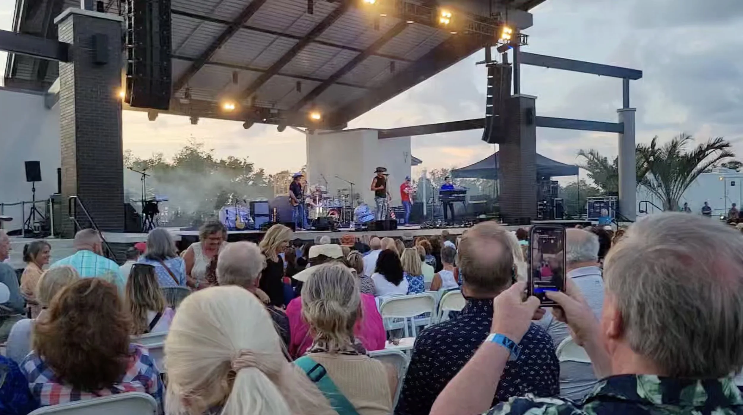 Outdoor concert with audience and band performing on stage under a pavilion.