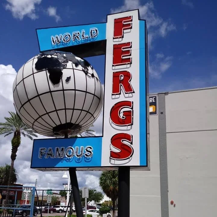 Outdoor sign with a globe and red letters spelling "FERGS," flanked by the words "WORLD FAMOUS" on a sunny day with palm trees in the background.