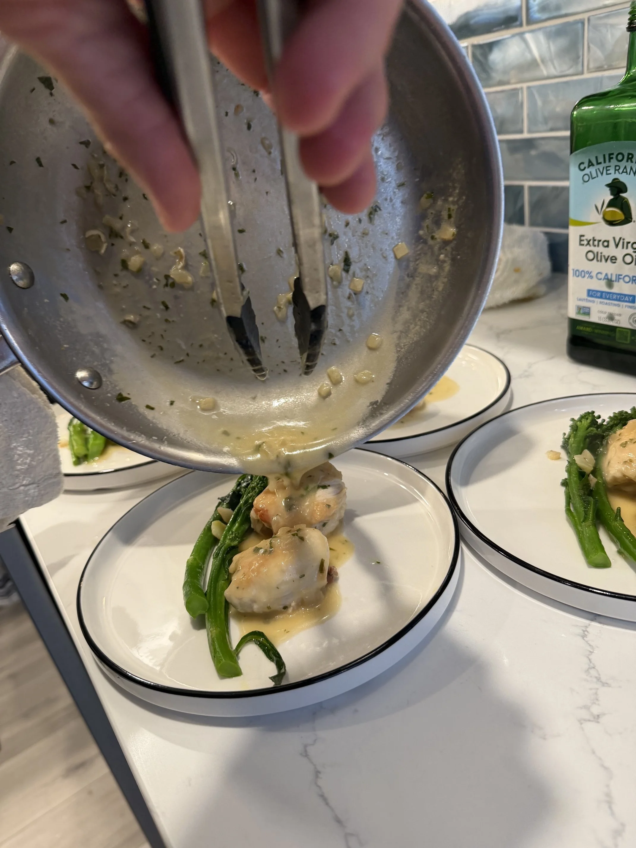 A person's hand holding tongs, serving a dish of cooked seafood and green beans with creamy sauce onto white plates.