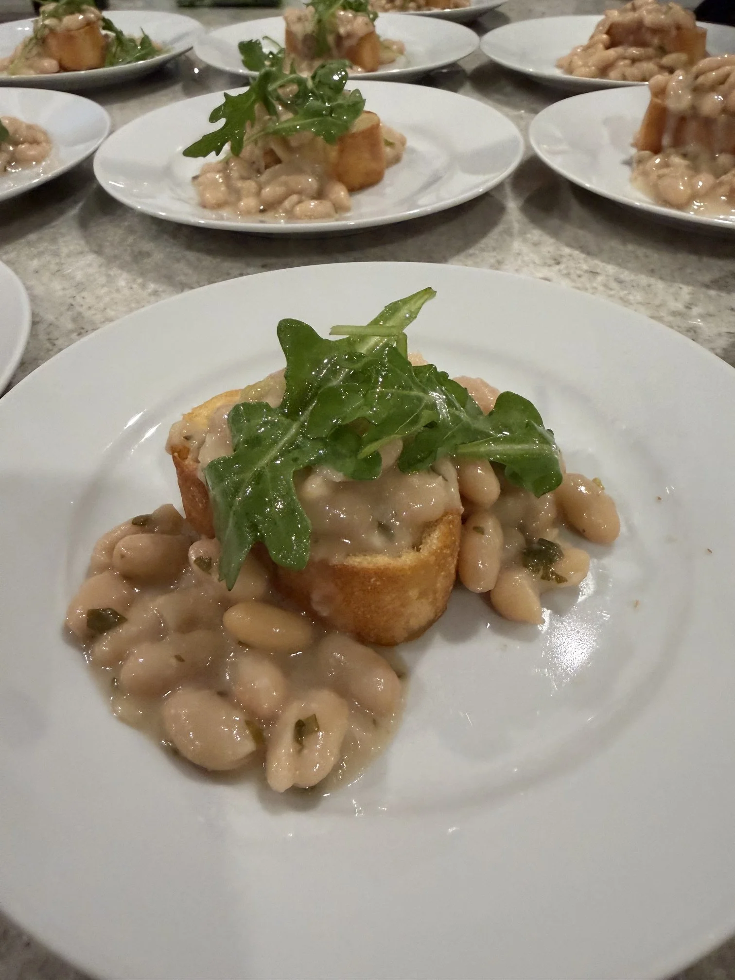 A white plate with a piece of toasted bread topped with white beans and a small green arugula leaf, with a creamy sauce plated by private chef Anthony Chiero of The Simmering Chef for an in-home dinner party in Newport, RI.