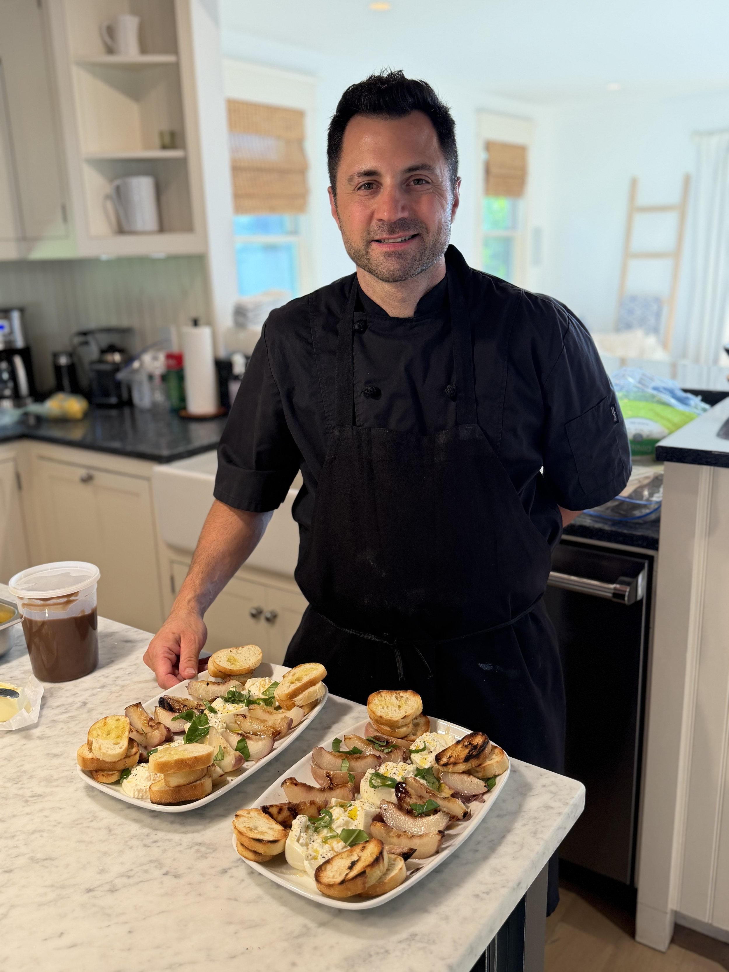 A man in a black chef's coat standing in a kitchen with three large plates of appetizers on a marble countertop, including sliced bread with toppings and garnishes, and a iced coffee drink on the side.