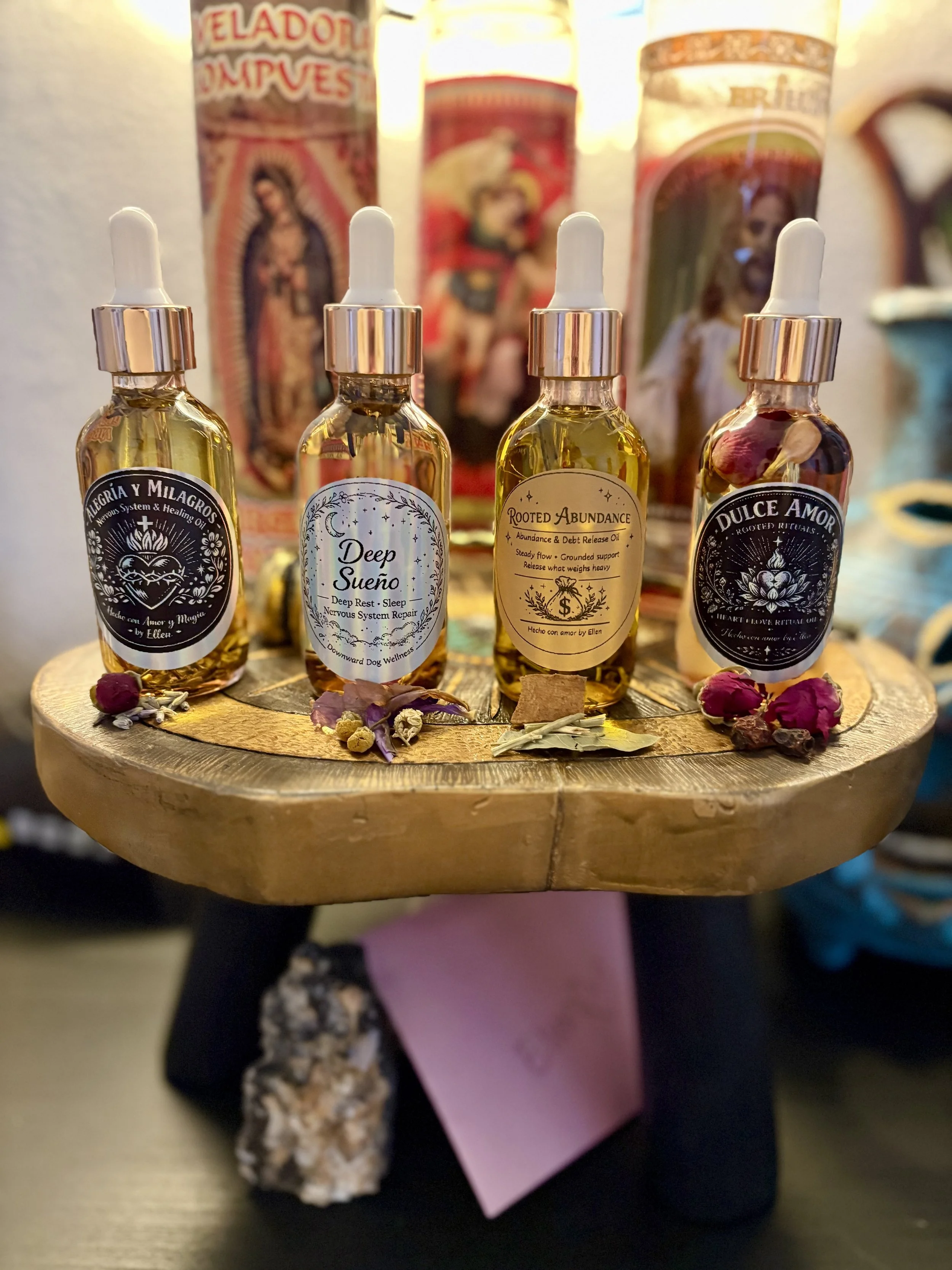 Four small bottles of essential oils with dropper caps are displayed on a wooden stand with flower petals and dried herbs around them. In the background, there are religious candles with images of saints.
