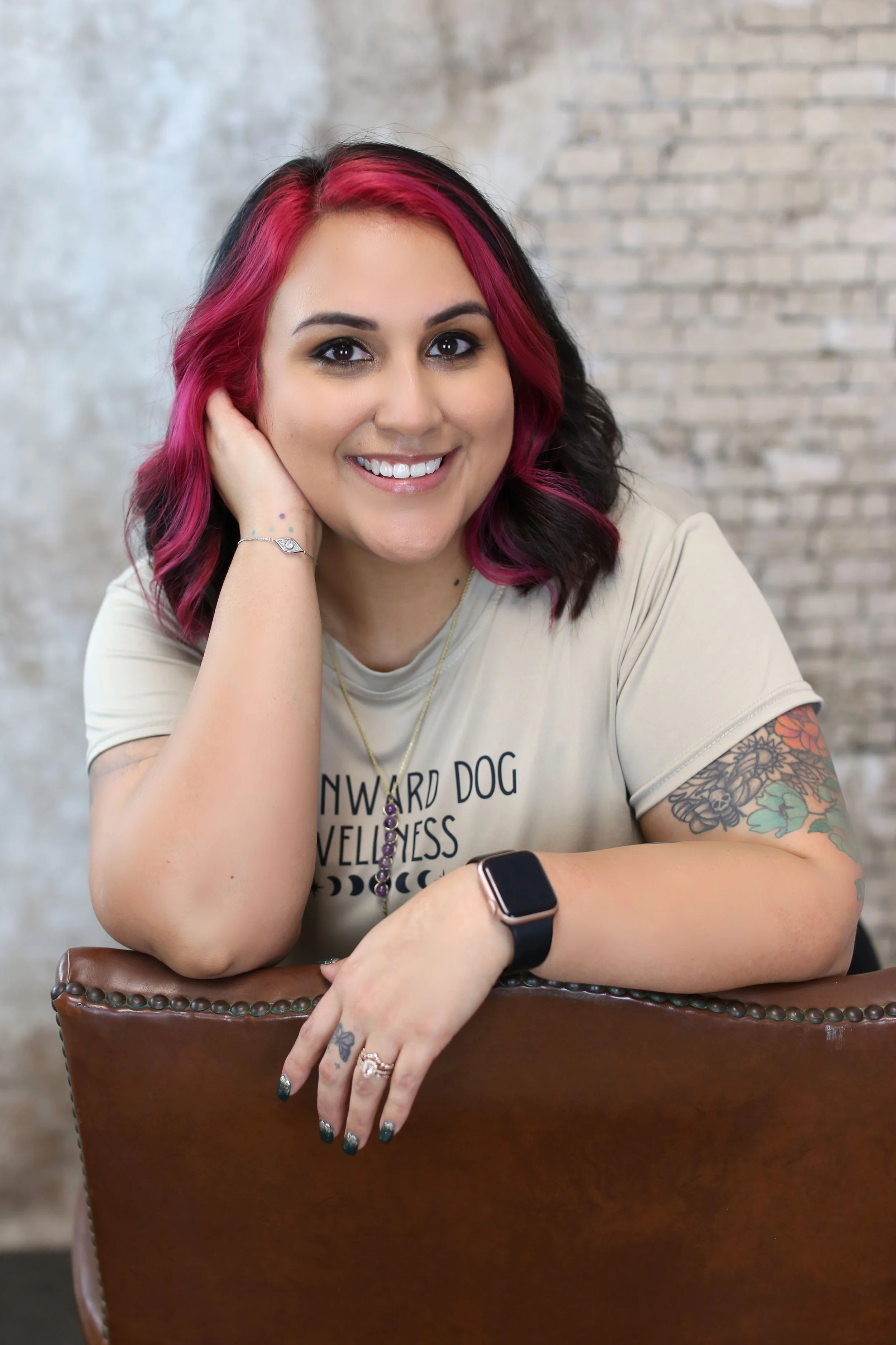A woman with shoulder-length dark hair with pink highlights, smiling and resting her head on her hand, sitting at a table in front of a brick wall.