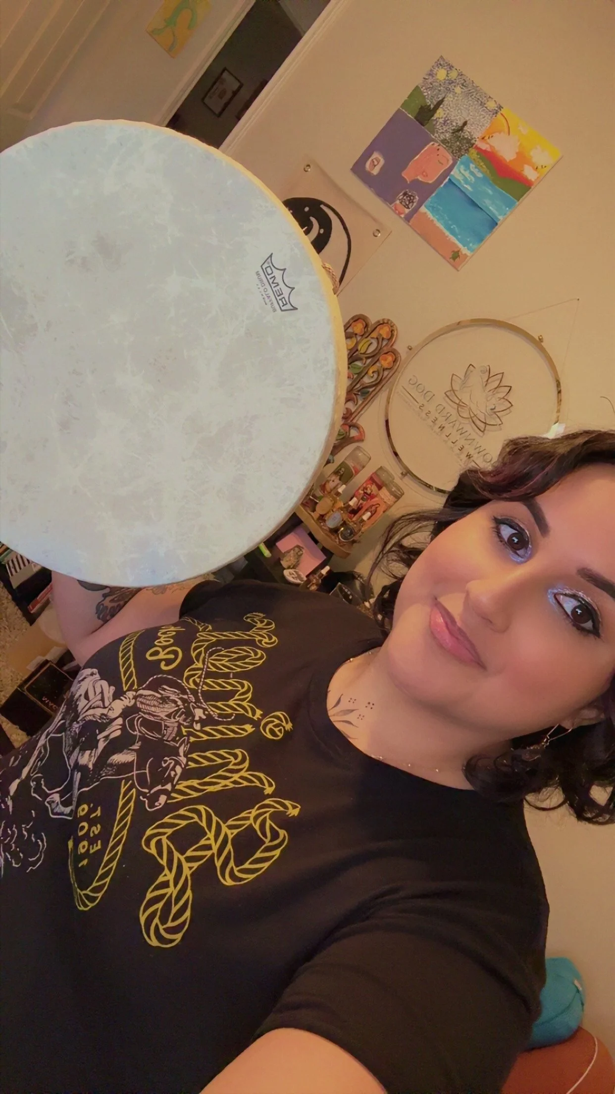 A woman taking a selfie in a room with colorful art and decor on the walls, holding a white circular lampshade in front of the camera, wearing a black t-shirt with yellow and white graphic design.