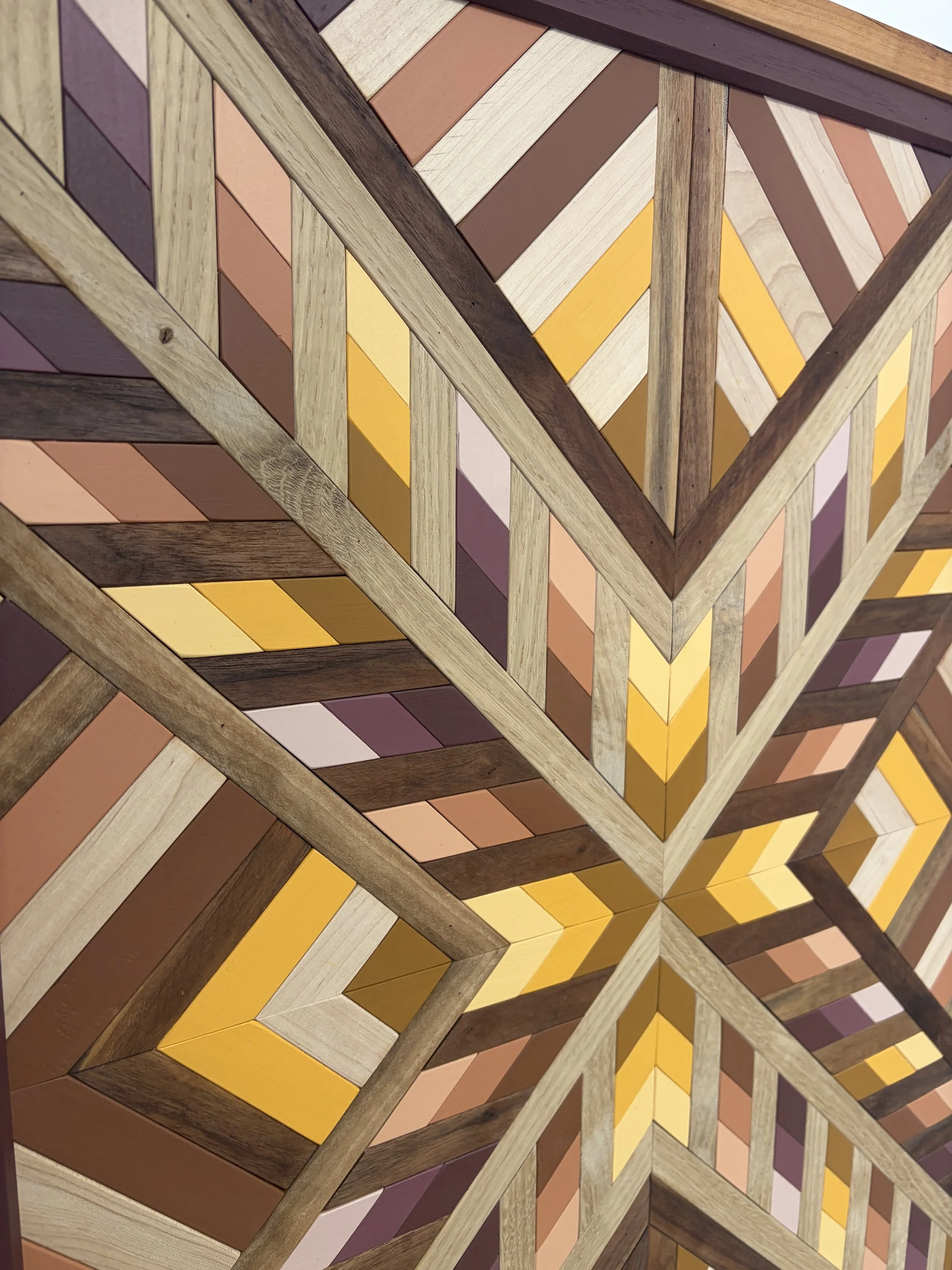 Close up view of a handcrafted framed wooden mosaic in several shades of pink, orange, and yellow paint with natural stains.