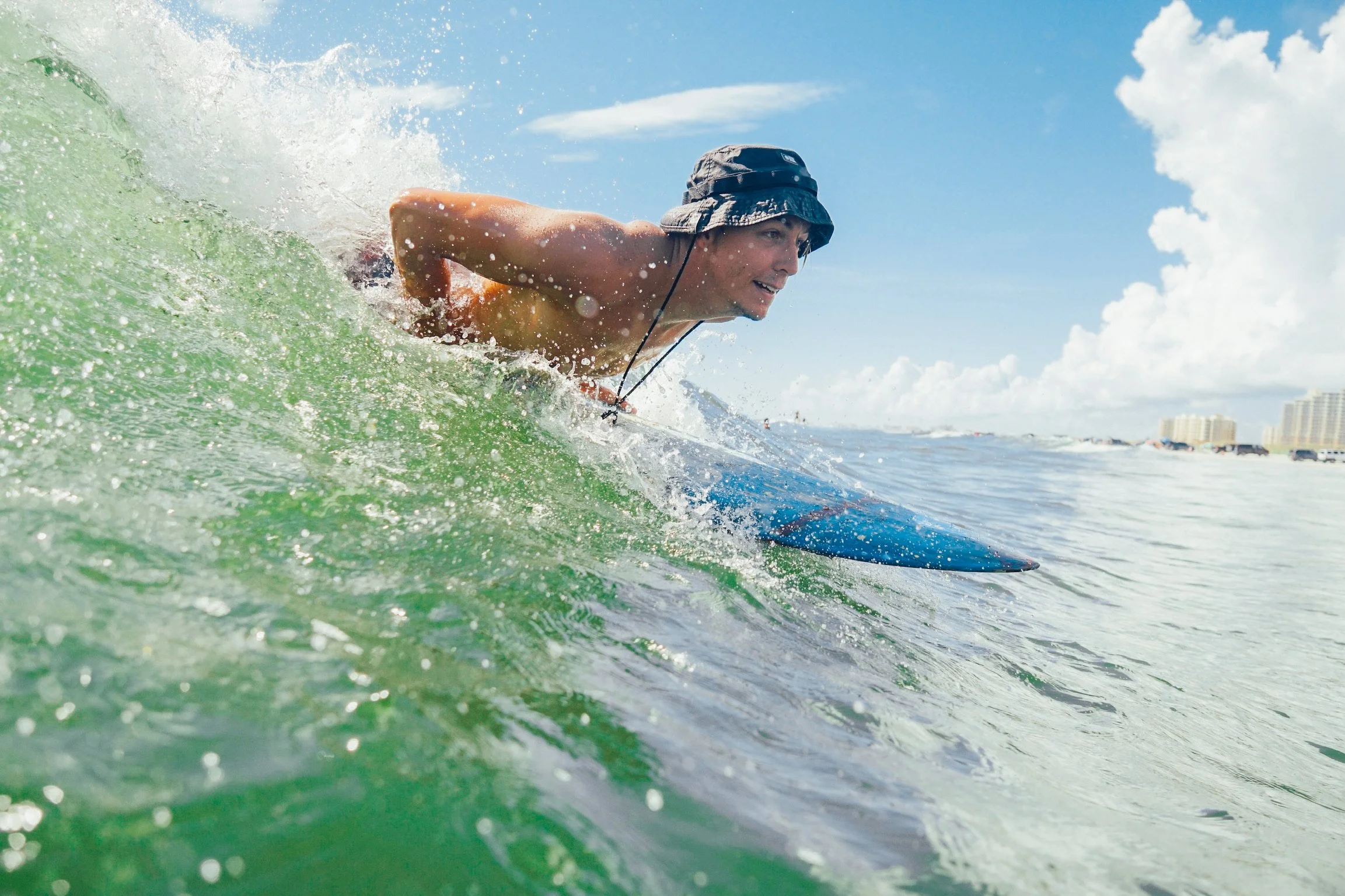 A person wearing a hat and no shirt surfing on a wave at the beach with buildings in the background and a blue sky with clouds.