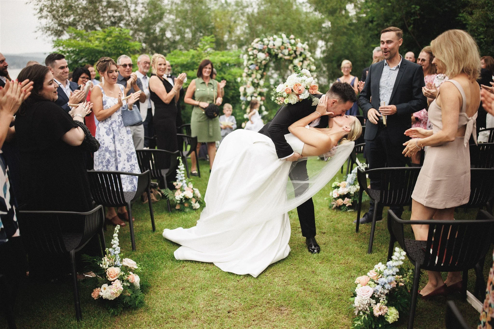 Un couple marié regarde l'un l'autre dans une rue urbaine ensoleillée, la mariée porte une robe blanche avec une longue traîne, tient un bouquet de fleurs roses et blanches, et porte une voile, tandis que le marié porte un costume noir avec une boutonnière blanche.