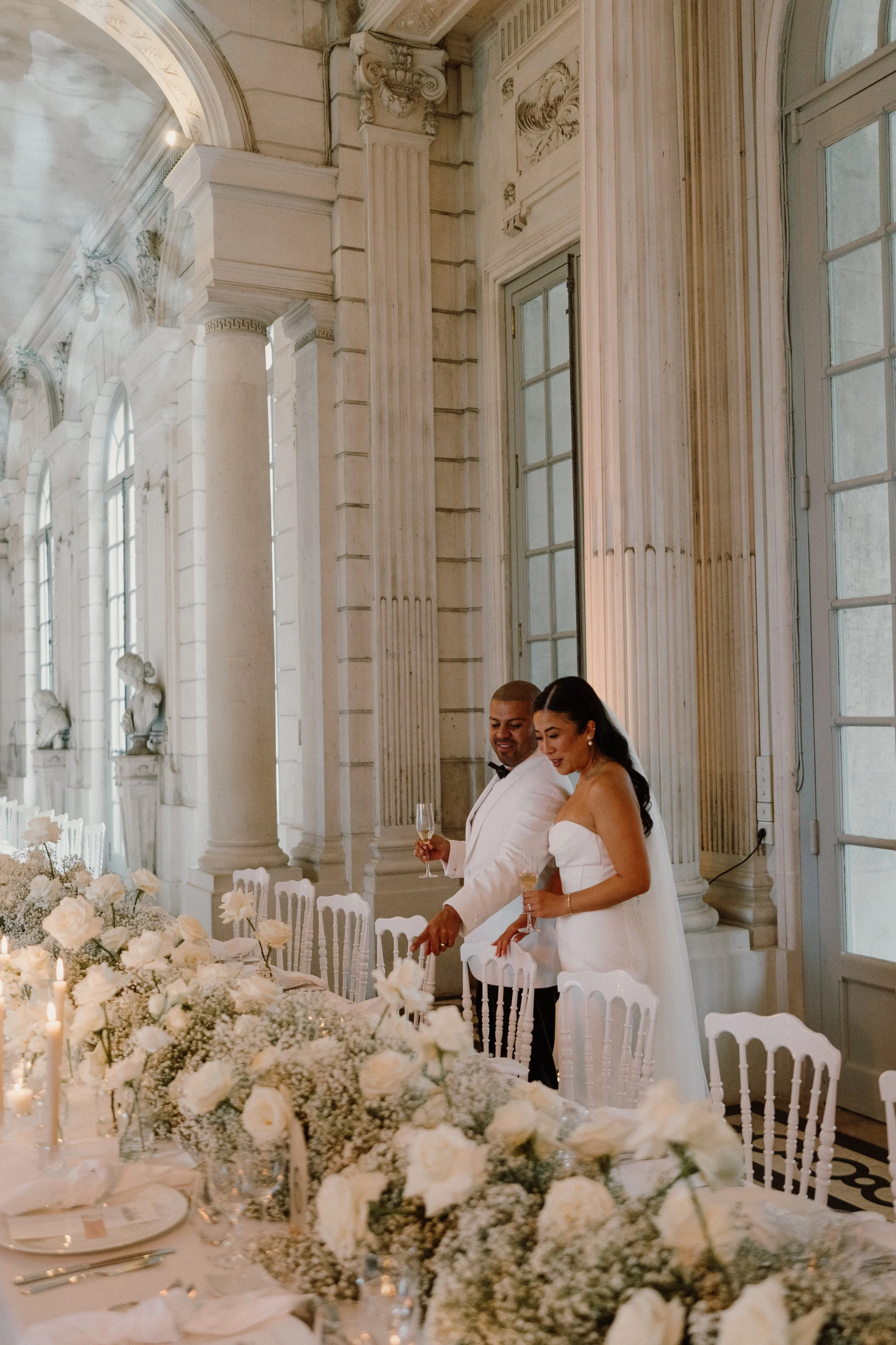 Deux personnes assises à une table lors d'un mariage, entourées de fleurs blanches et de bougies, dans une pièce avec des fenêtres en arrière-plan.