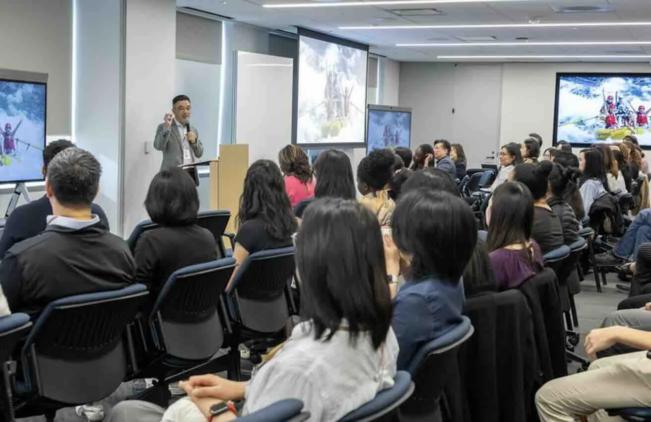 A man giving a presentation to a large audience in a conference room, with slides displayed on two monitors behind him.