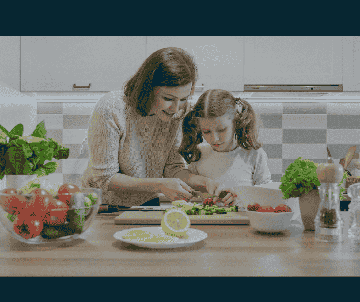 A woman and young girl prepare food together in a modern kitchen, slicing vegetables on a cutting board surrounded by fresh produce and kitchen items.