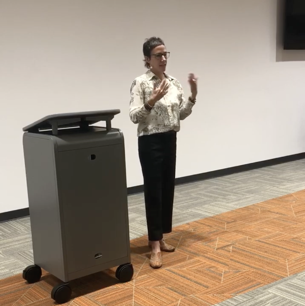 A woman giving a presentation in an office or conference room, standing near a gray podium or audiovisual equipment, with a large blank wall behind her.
