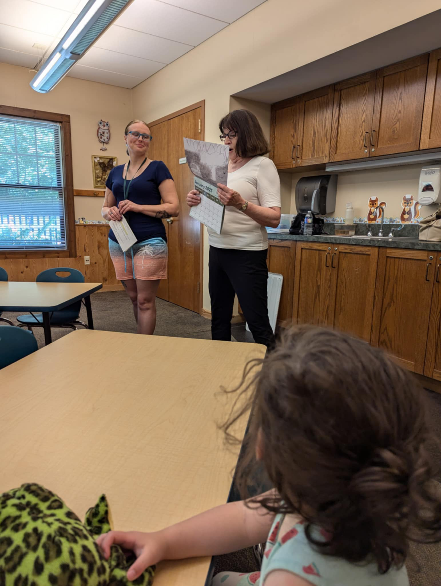 Two women standing in a room, one holding a calendar and the other with a piece of paper, while a child with curly hair and another with leopard print clothing are seated at a table in front of them.