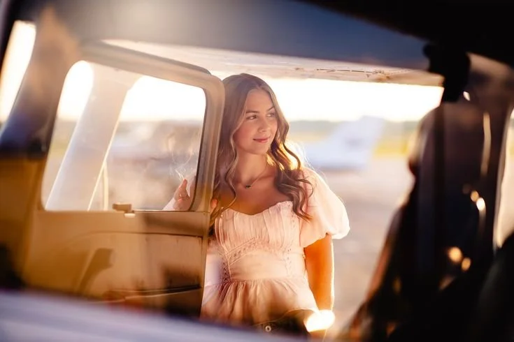 Stand in the open doorway and lightly hold the frame. Look out toward the ramp first, like you’re about to start your flight, then give me a soft glance back. Look out, take a breath, then slowly turn your eyes back to me