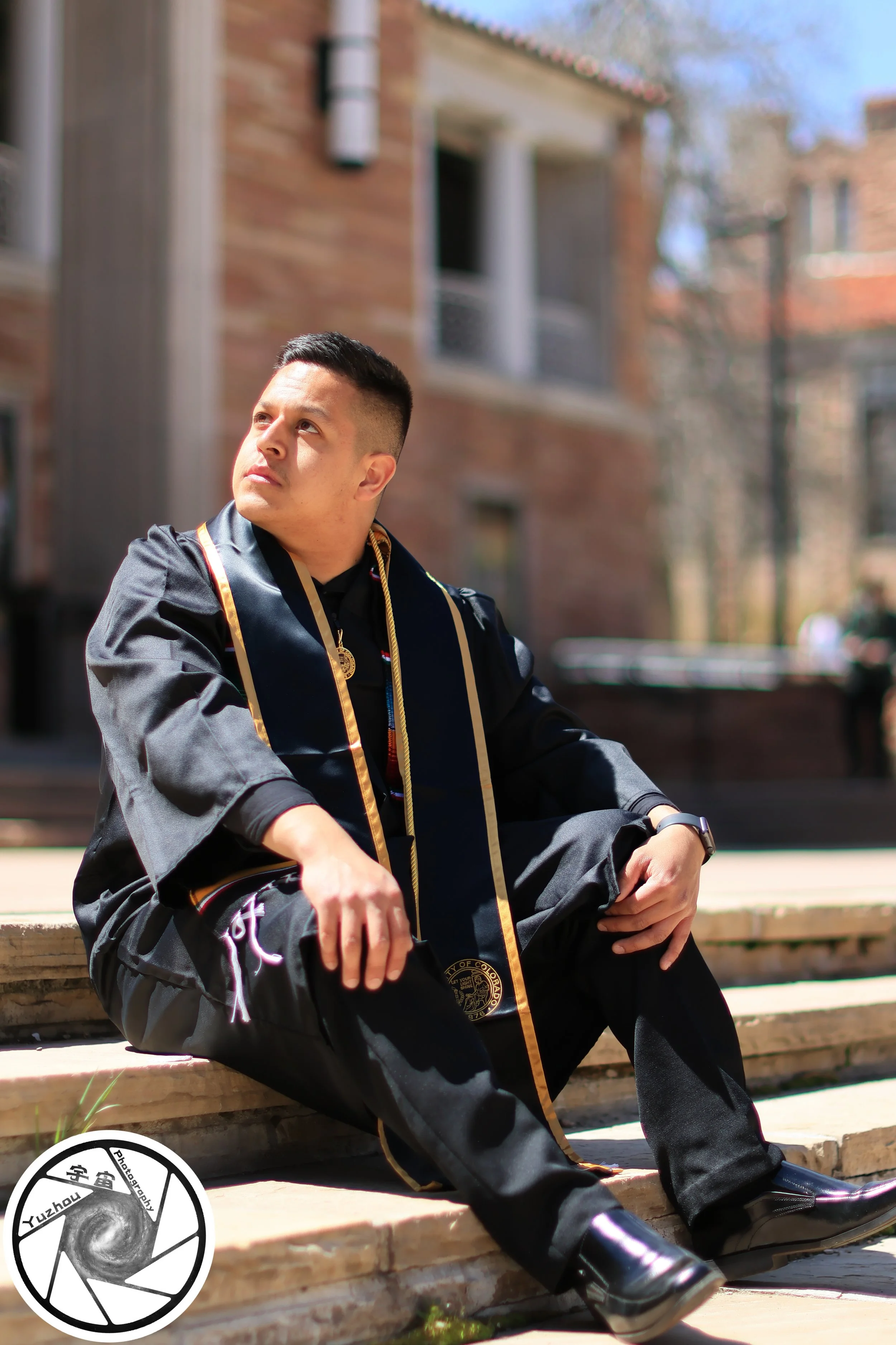 Young man in graduation gown and cap sitting on steps outdoors, looking thoughtfully into distance.