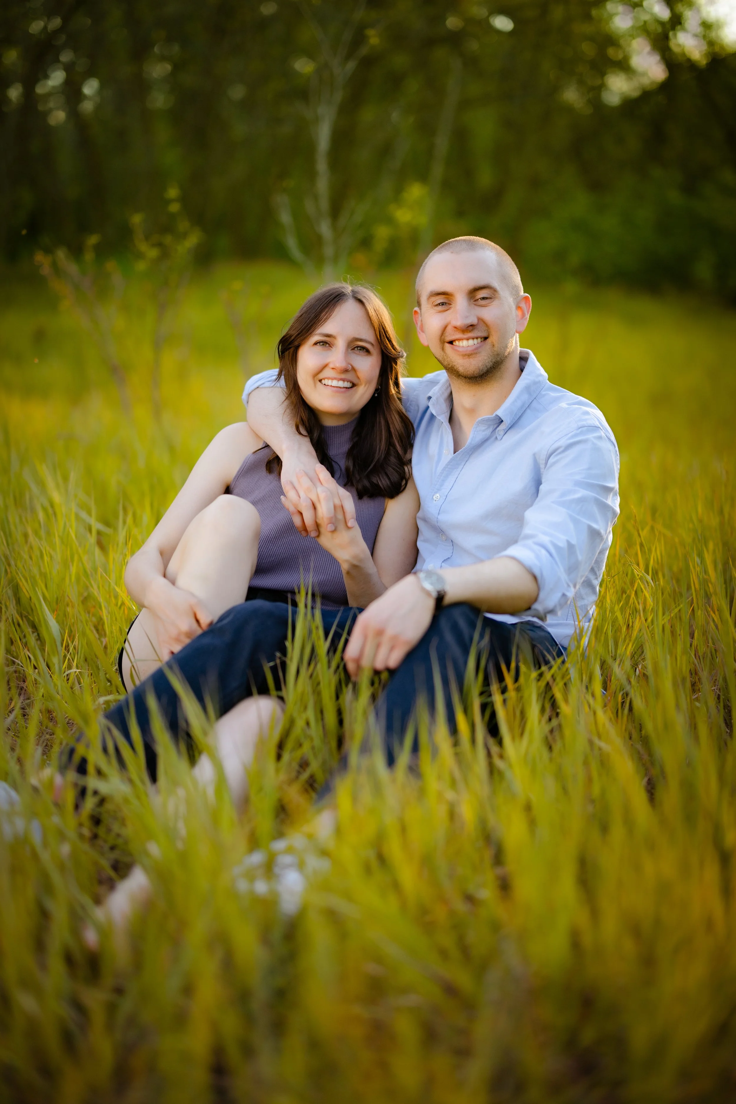 A smiling couple sitting in a grassy field with trees in the background during sunset.