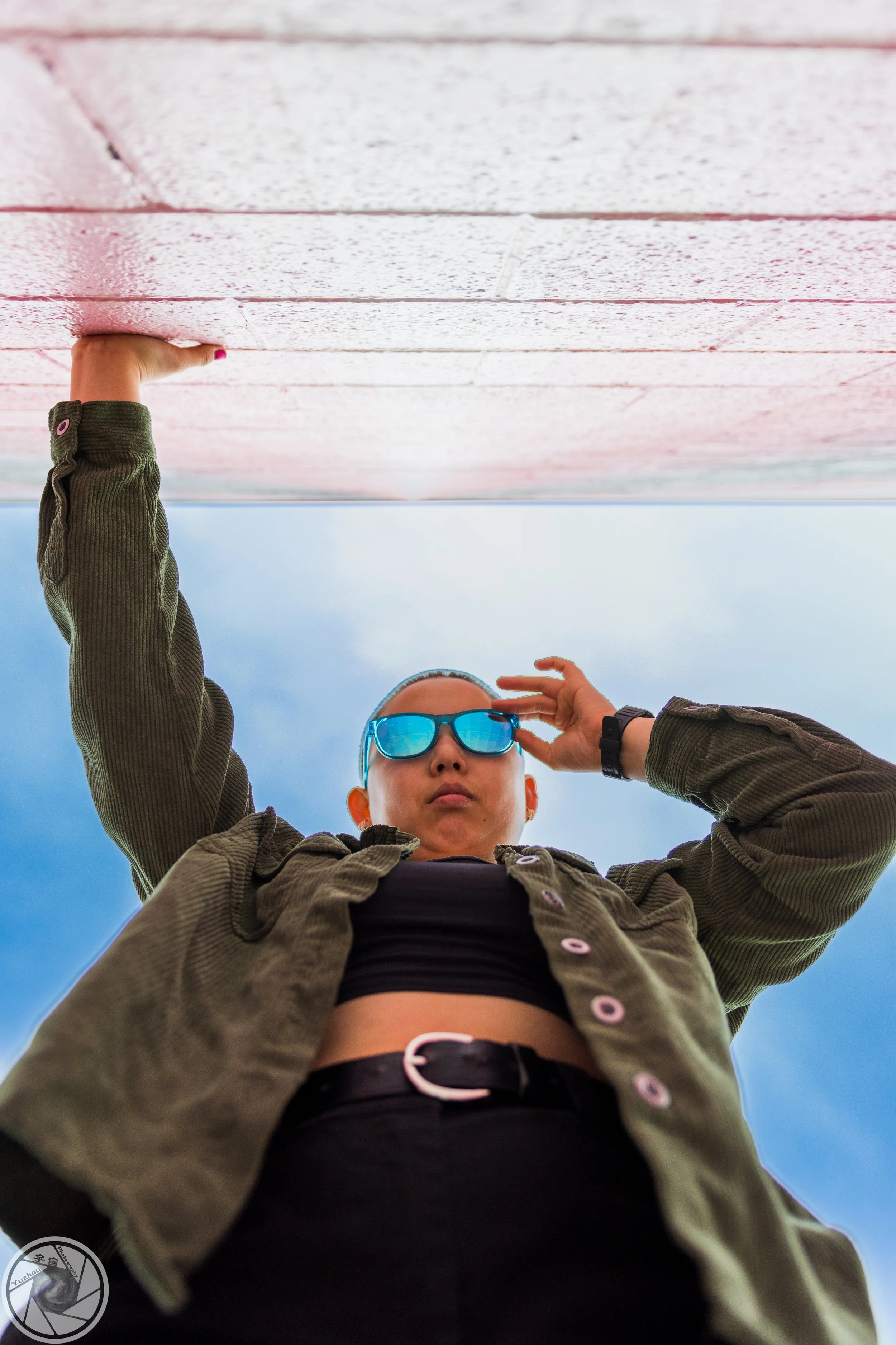 Low-angle shot of a woman with blue sunglasses, wearing an olive green jacket and black crop top, looking down at the camera from beneath a concrete ledge against a cloudy sky.