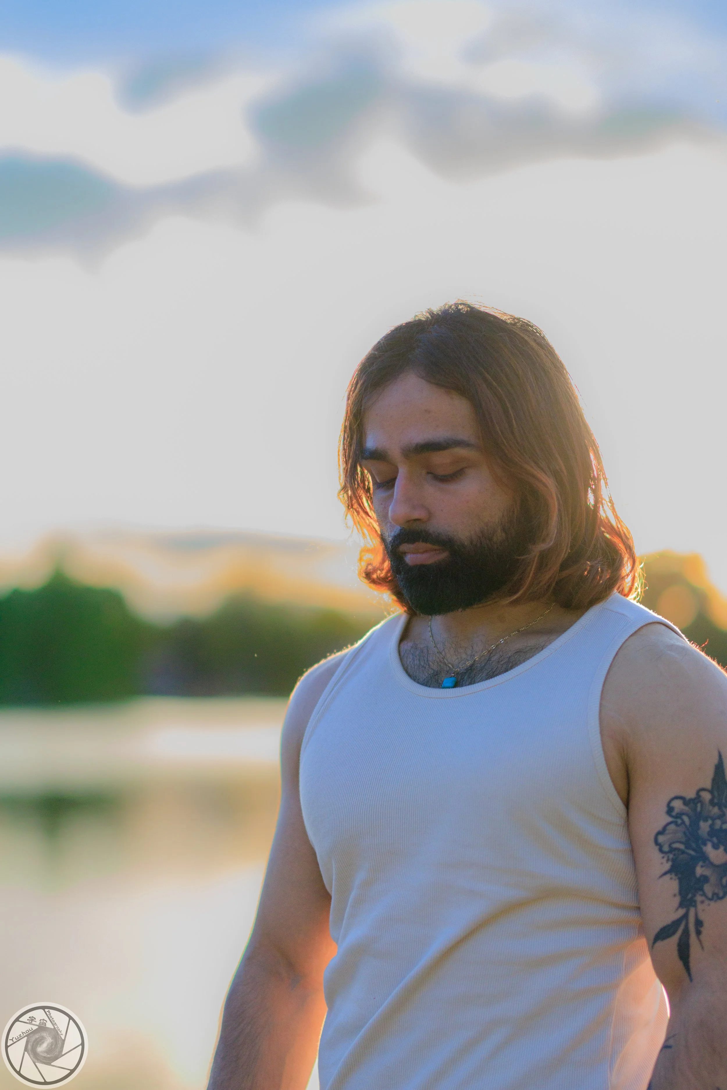 A man with long hair and a beard wearing a white tank top and a tattoo on his upper arm stands outdoors near a body of water during sunset, with his eyes closed and a calm expression.