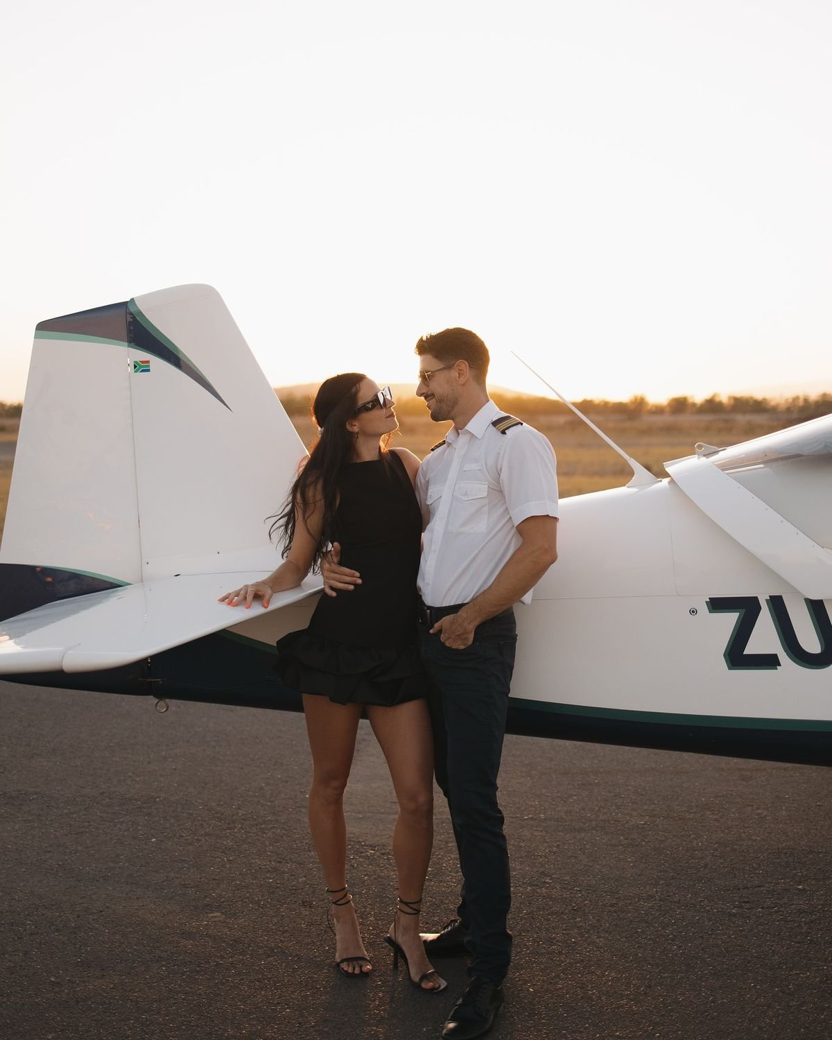 Stand near the tail section and place one hand lightly on the aircraft. Turn your body toward the sunset, then look back toward me with a confident, calm expression.”
