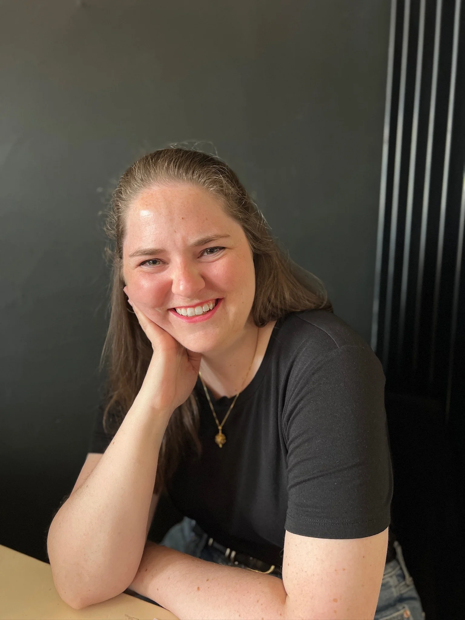 Close-up of a smiling woman with long brown hair, resting her chin on her hand, wearing a black shirt and a gold necklace, sitting at a desk against a dark background with vertical black and white stripes.