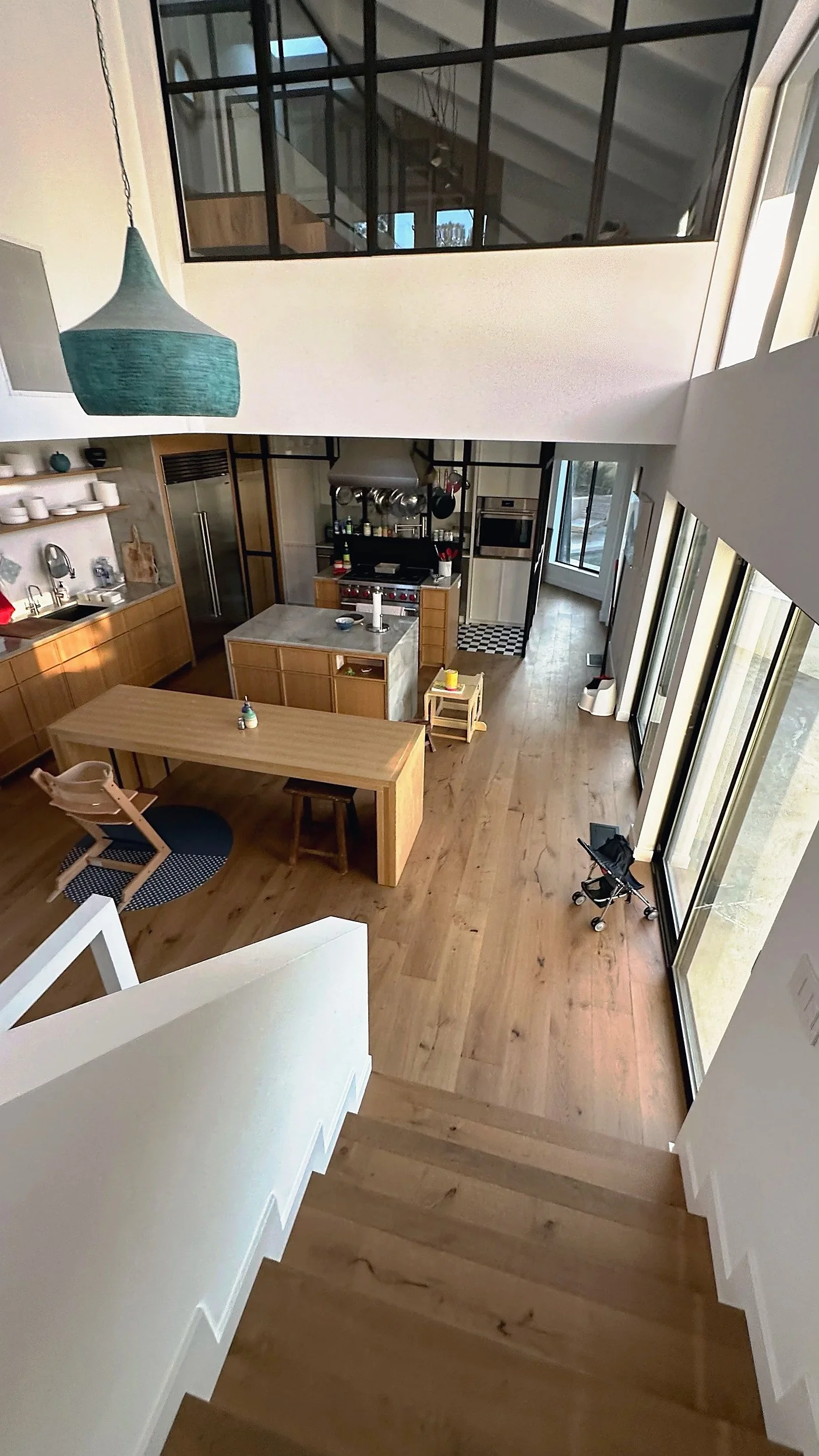 Modern kitchen with wooden cabinets, island, and dining table viewed from upstairs. Large windows, pendant light, and a small staircase on hardwood floor.