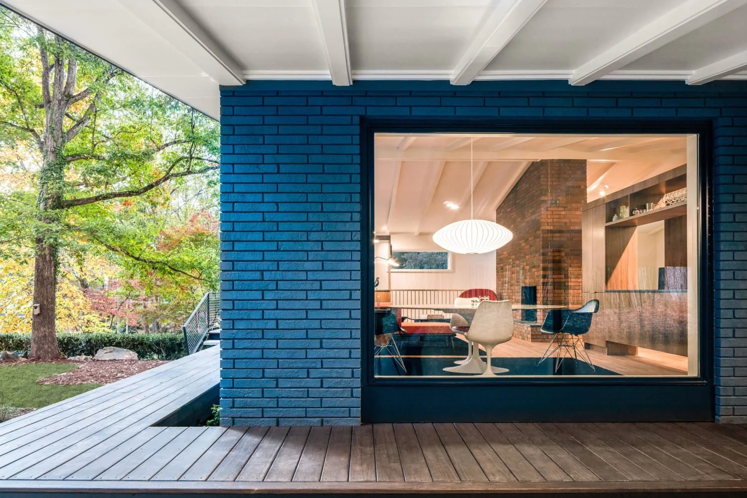 Contemporary home exterior with blue brick wall, large window showing modern interior seating, pendant light, and surrounded by lush trees.