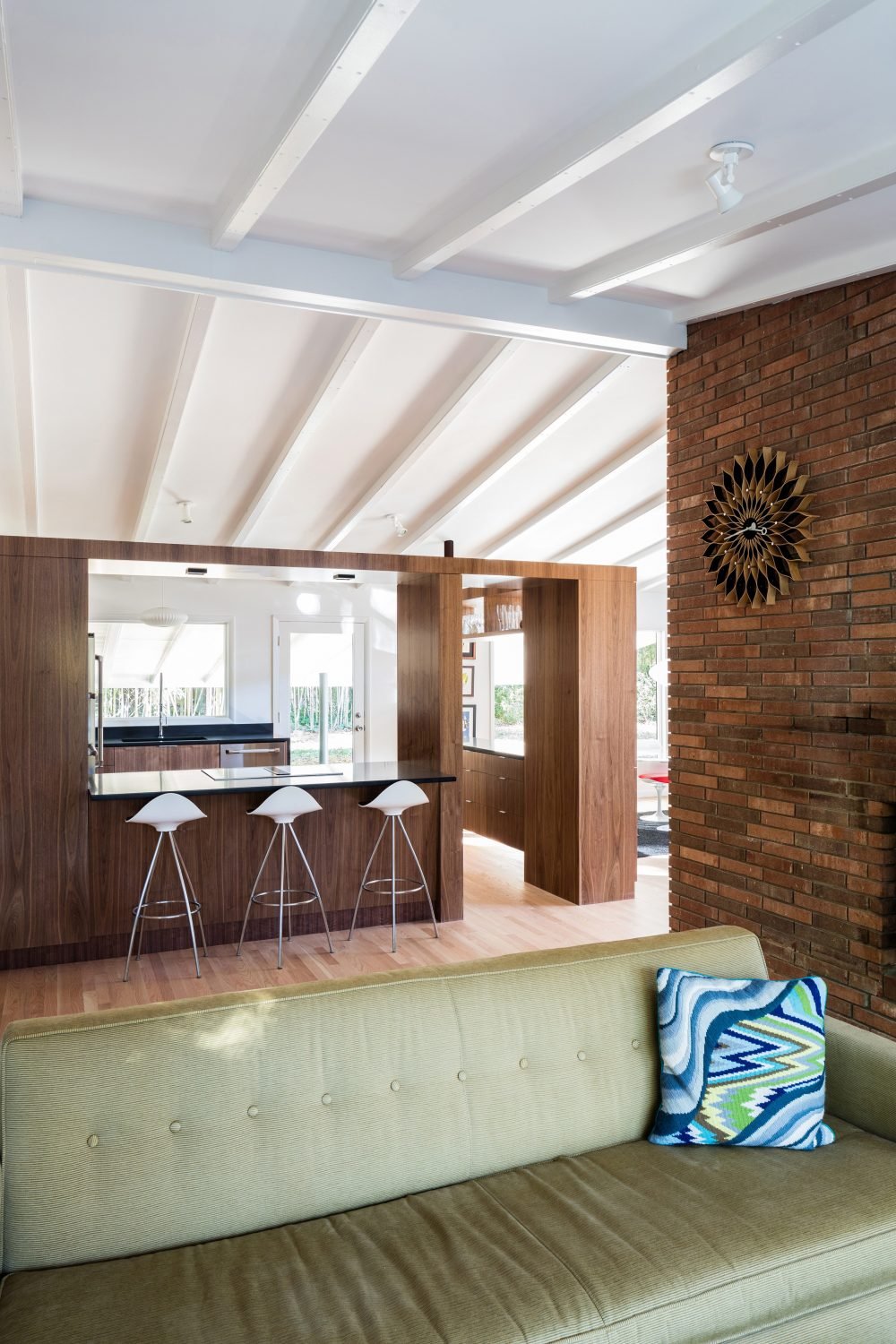 Modern living room with green sofa, colorful cushion, wooden kitchen counter with bar stools, brick wall with decorative clock.