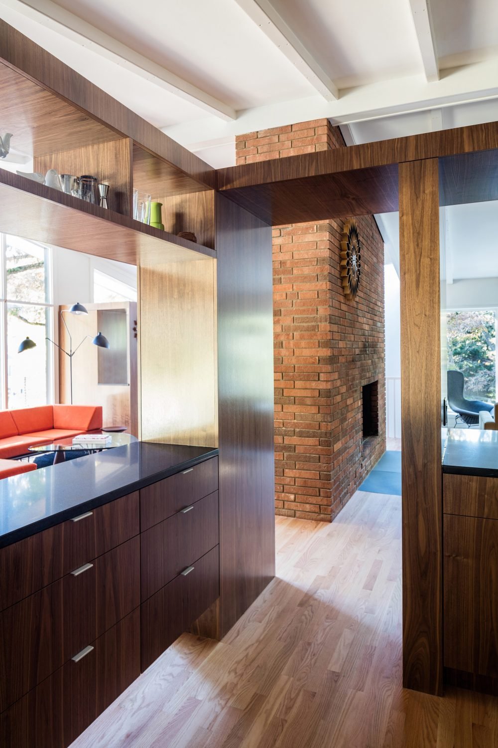 Modern interior with wooden cabinets, brick fireplace, red couch, and floor-to-ceiling windows. The room is bright and features contemporary decor, including a decorative wall clock.