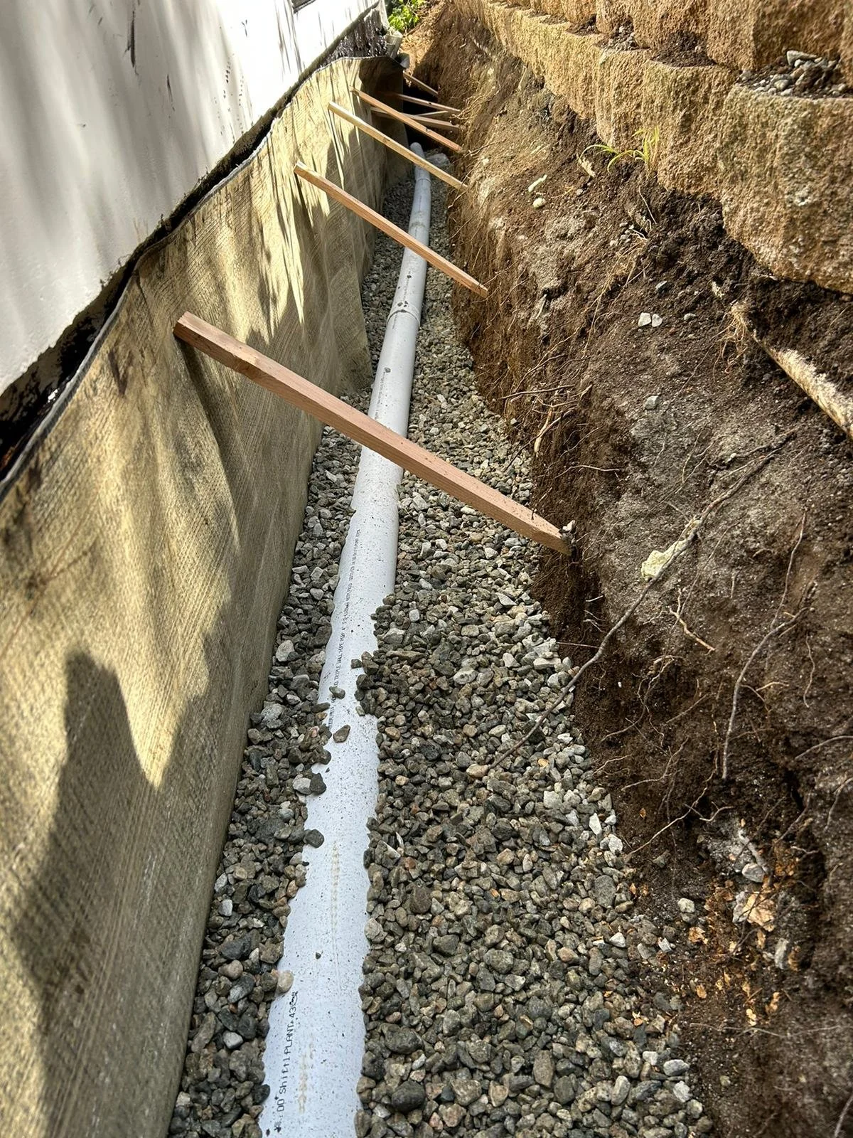 Trench with drainage pipe being installed, surrounded by gravel, against a retaining wall and building foundation.