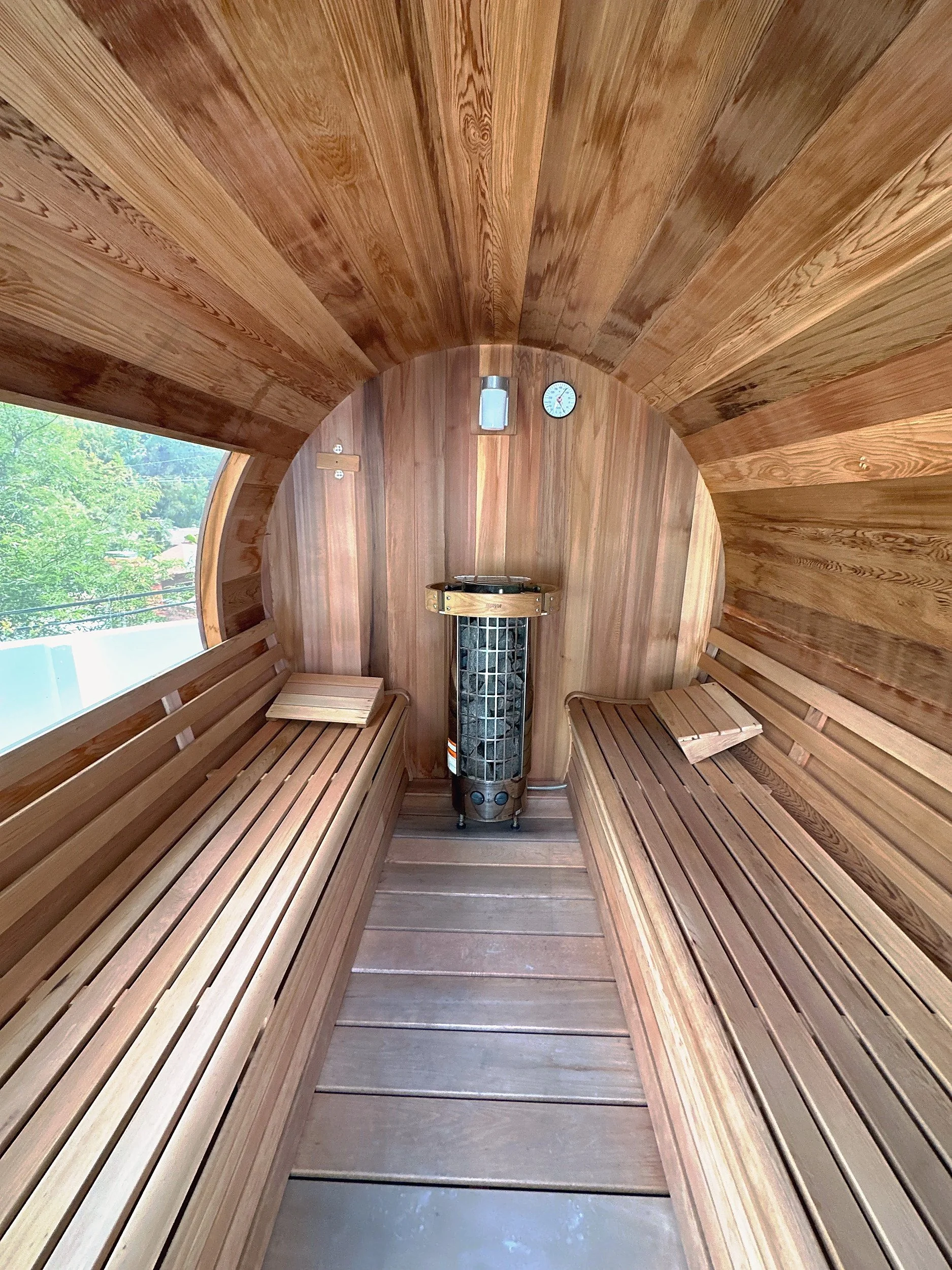 Interior of a wooden barrel sauna with benches, a metal heater, and a wall clock.