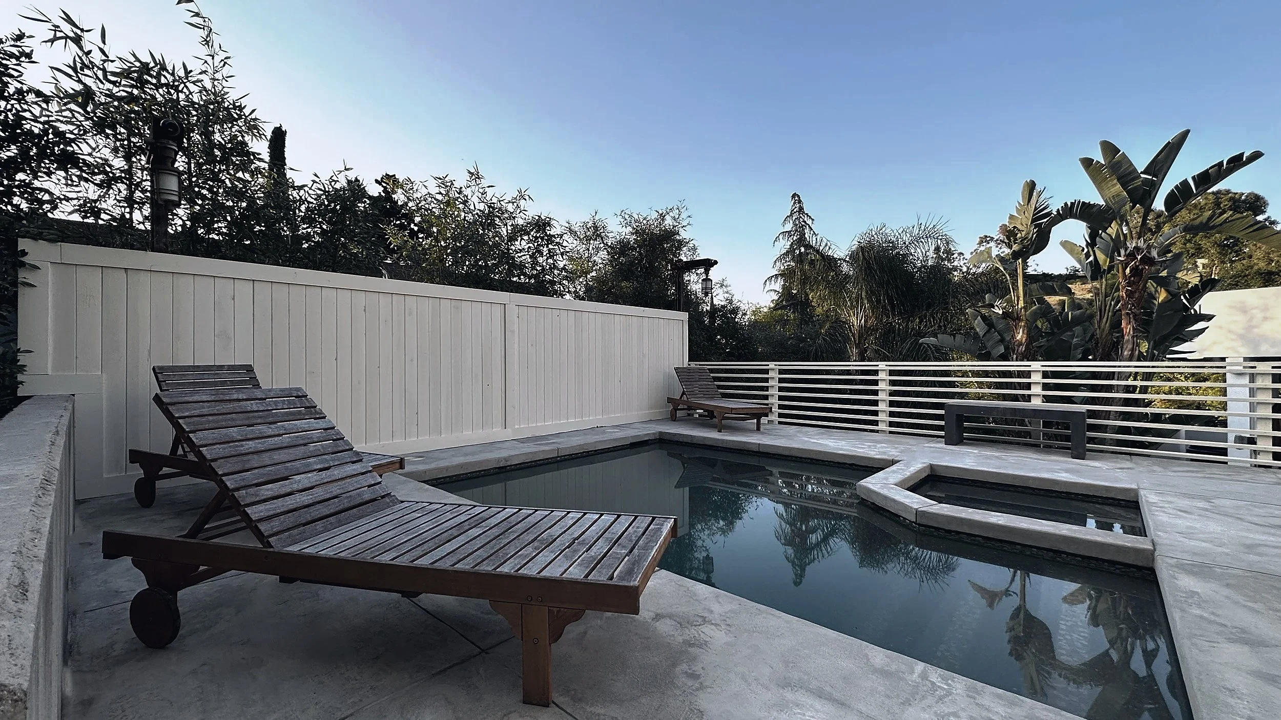 Pool and spa set in a concrete terrace, with wooden loungers and a painted board fence.
