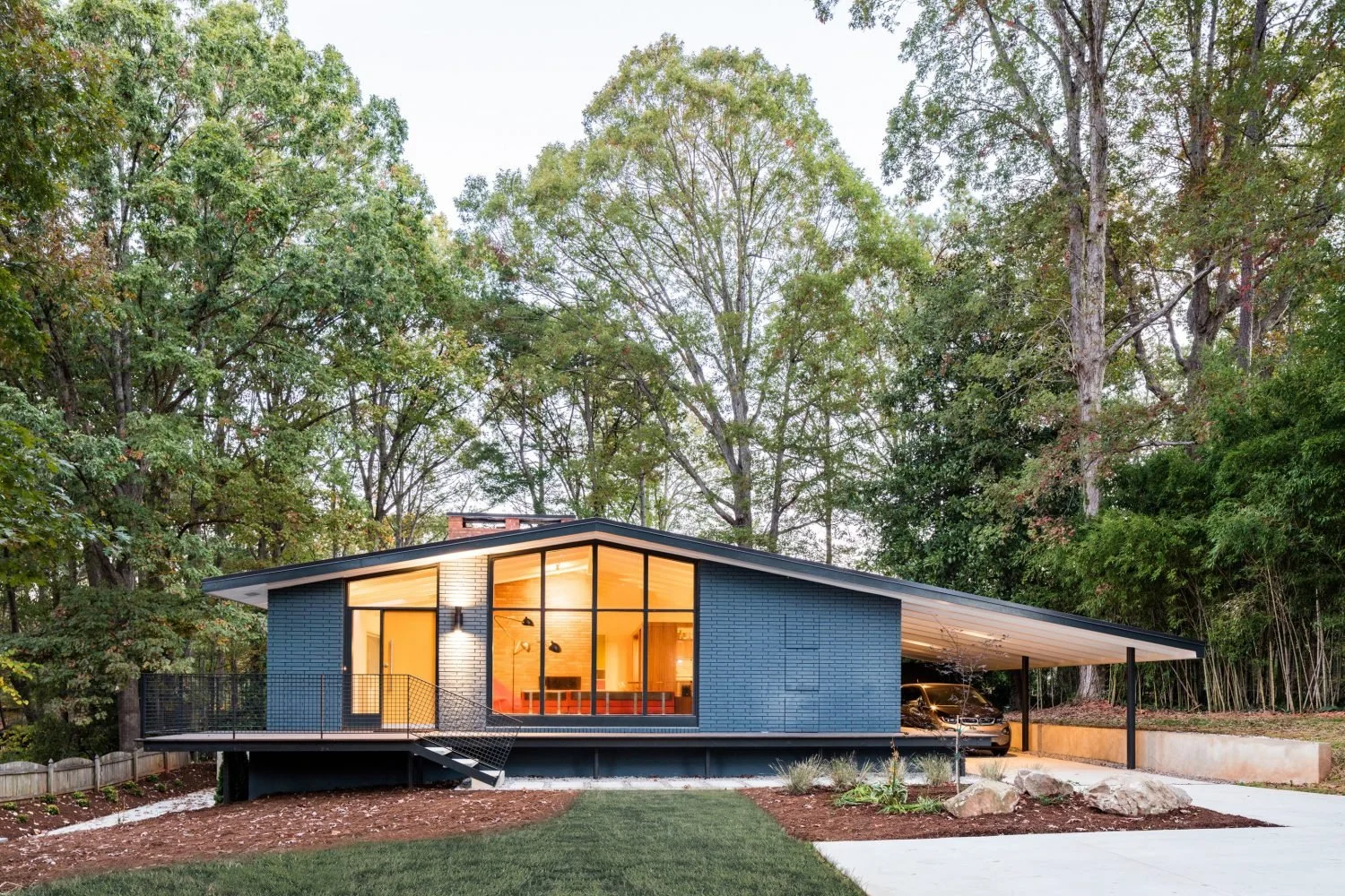 Modern blue house with large windows, surrounded by trees, featuring a sloped roof and a carport.