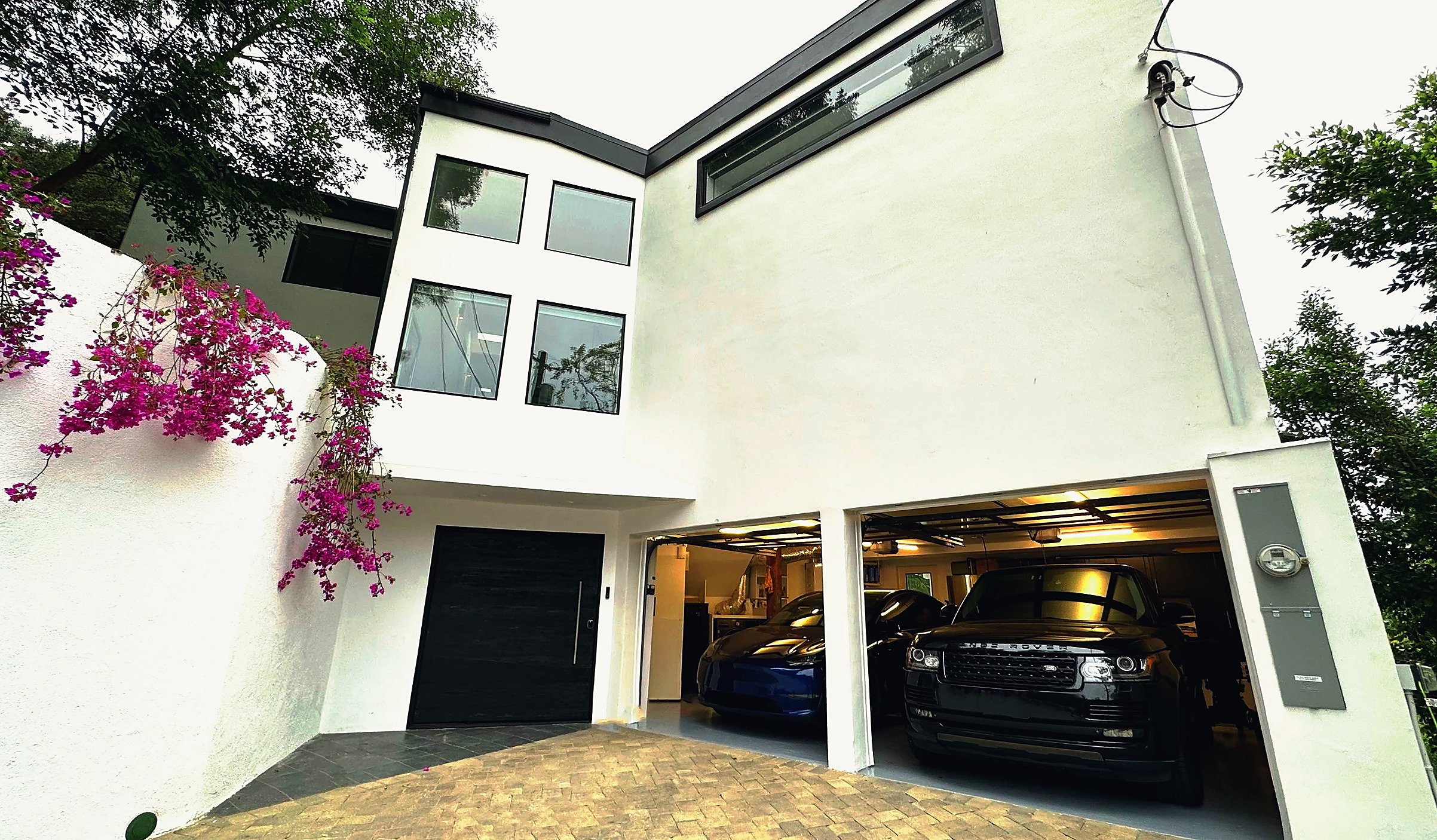 Modern white house with large windows and a two-car garage containing a blue car and a black SUV. Pink flowers hang over a white wall beside the driveway.