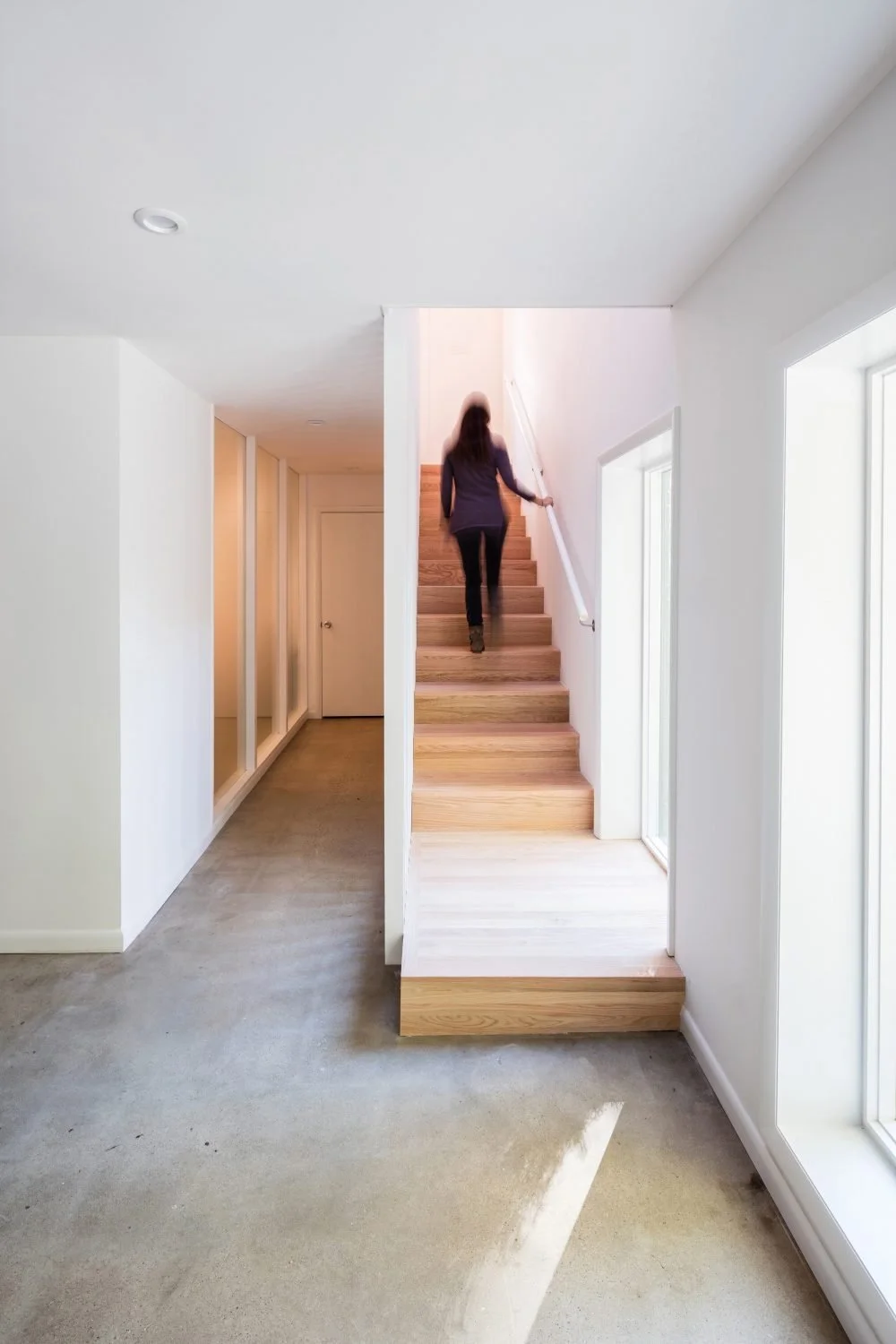 Person walking up stairs in modern, minimalist hallway with white walls and wood steps.