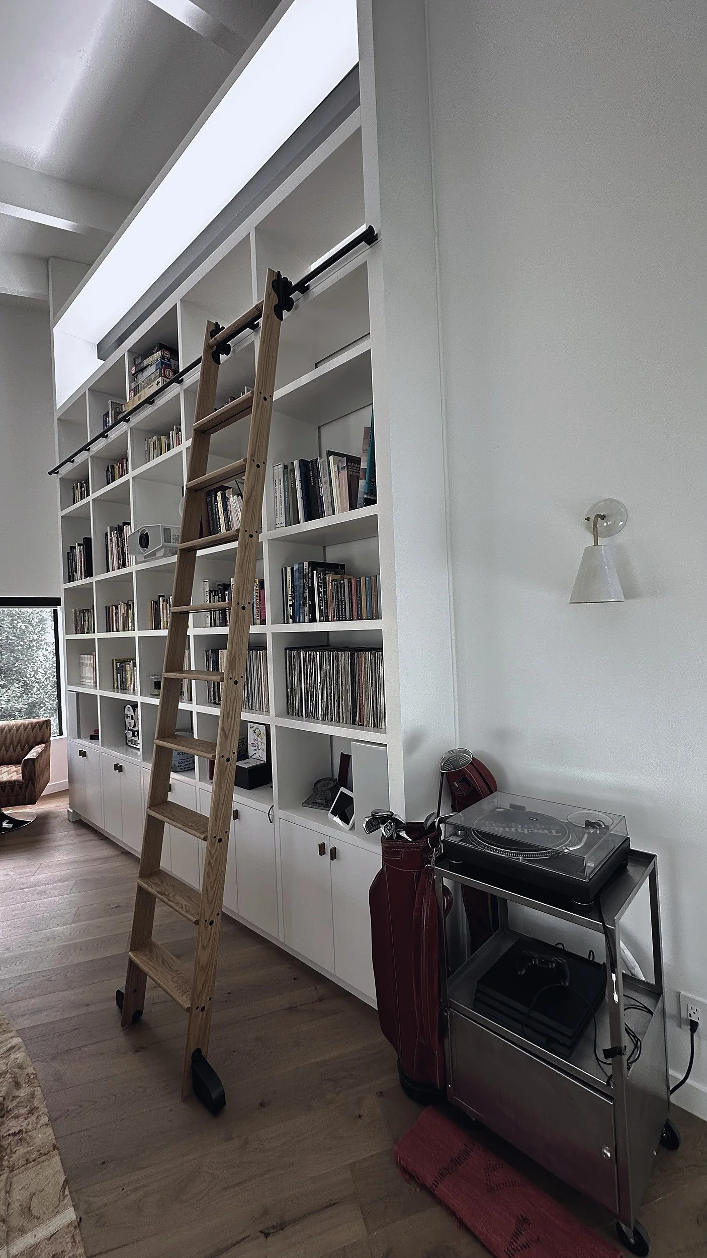 Double-height white built-in shelving wall holding books and records, with an oak rolling ladder.