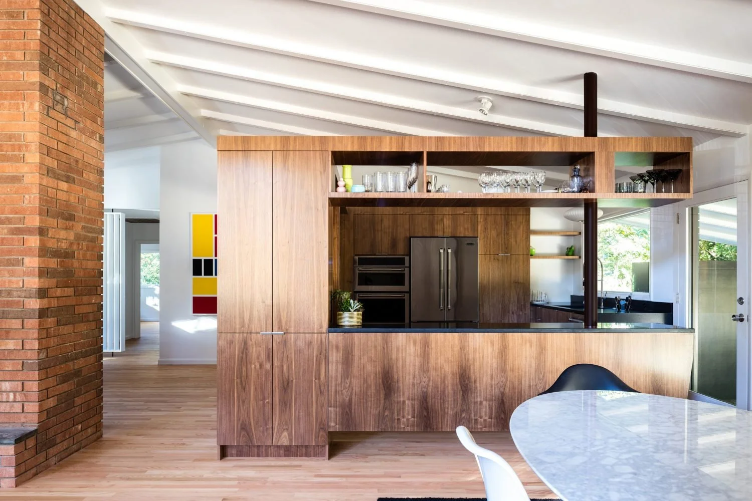 Modern kitchen with wooden cabinetry, stainless steel appliances, open shelving with glassware, and a marble dining table. Exposed brick wall on the side and a color block artwork on the interior wall.