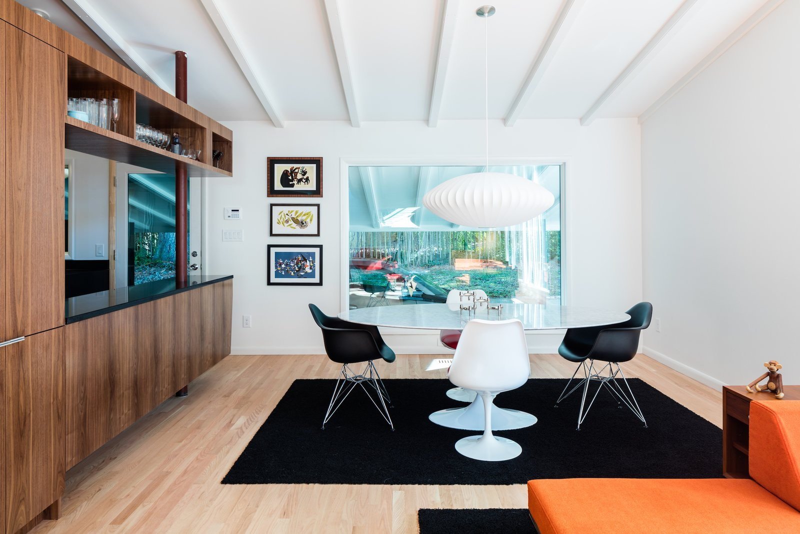 Modern dining room with Eames chairs, white pendant light, and wooden cabinetry. Large window overlooks garden. Art prints on wall, black rug on light wood floor, and an orange sofa in the foreground.