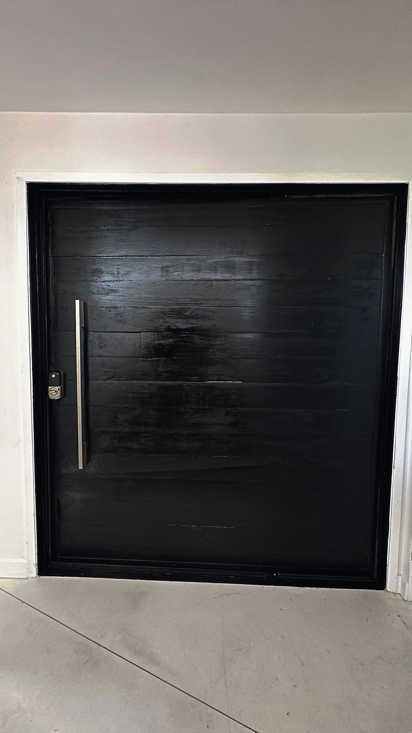Large black wooden door with a vertical metal handle and a digital lock on the left side, set in a white wall.