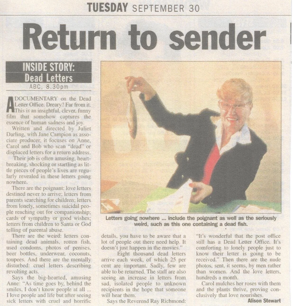 A woman with glasses, smiling, wearing a black blazer and white shirt, holding a dead fish by its tail over a desk full of papers, in a cheerful manner.
