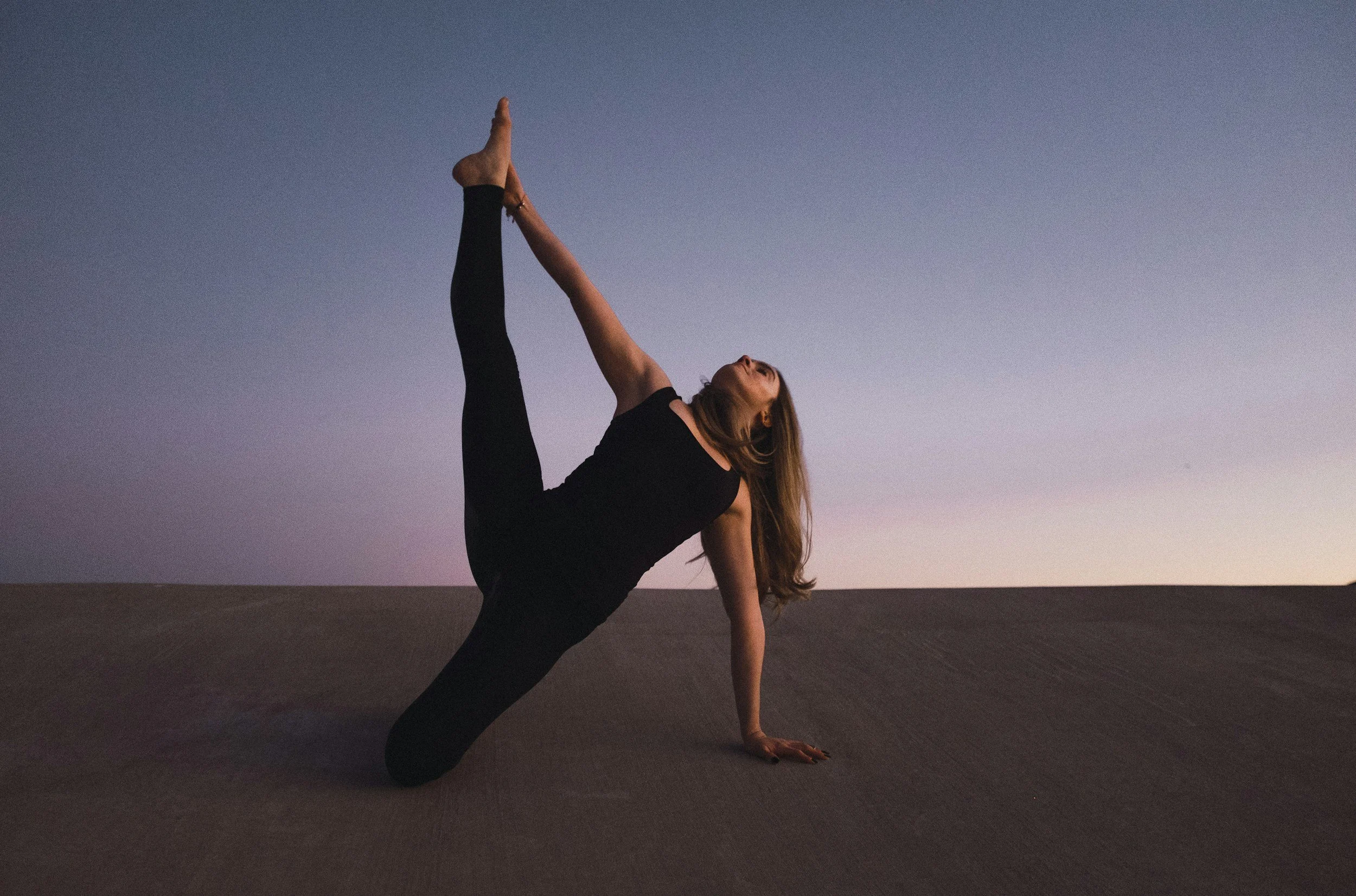 Una mujer haciendo yoga en postura en el desierto al atardecer, en posición de triángulo, con el cielo en tonos morados y anaranjados.
