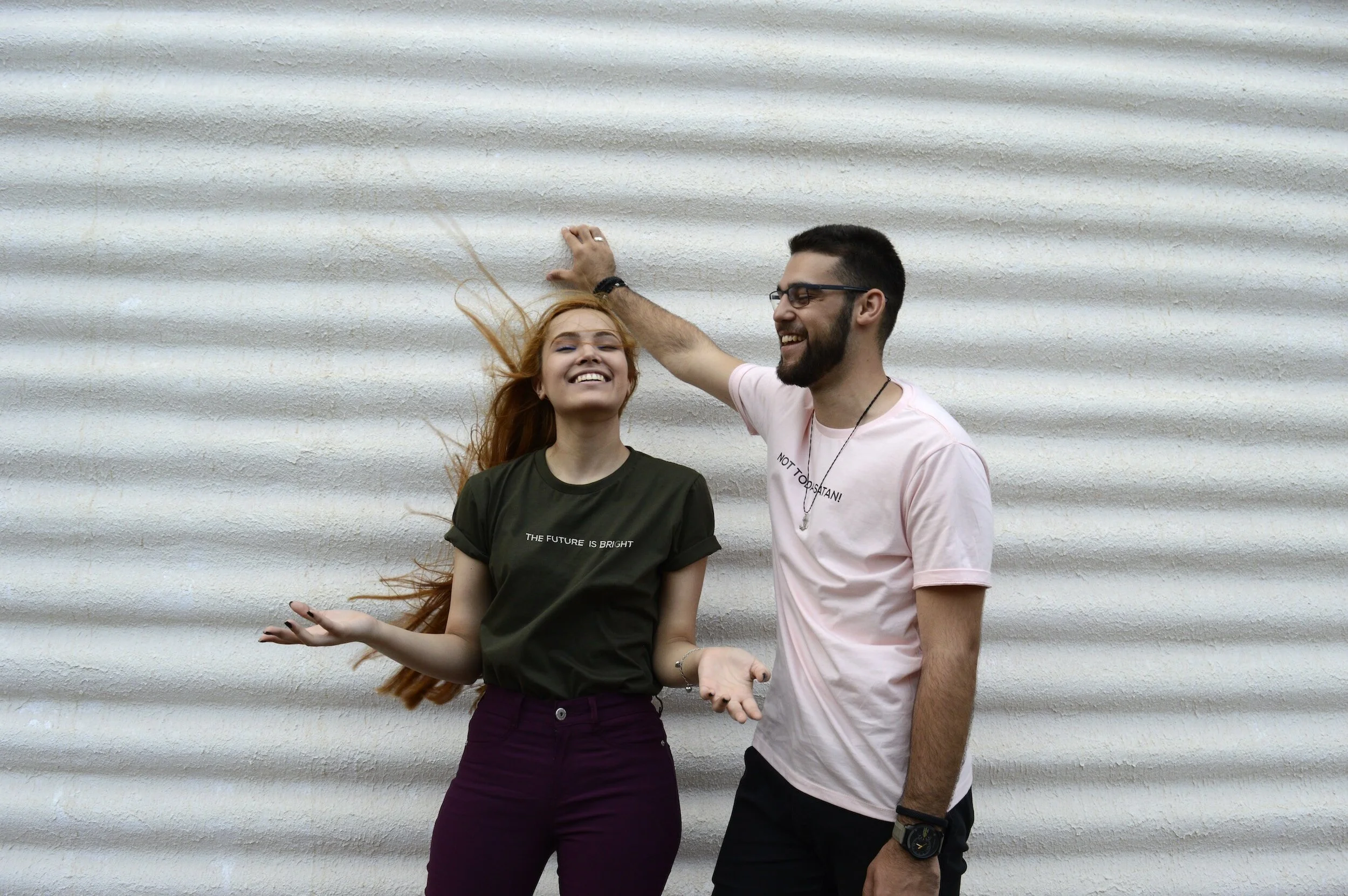 A woman with long hair in a green shirt and a man with a beard in a pink shirt standing against a corrugated metal wall, both smiling.
