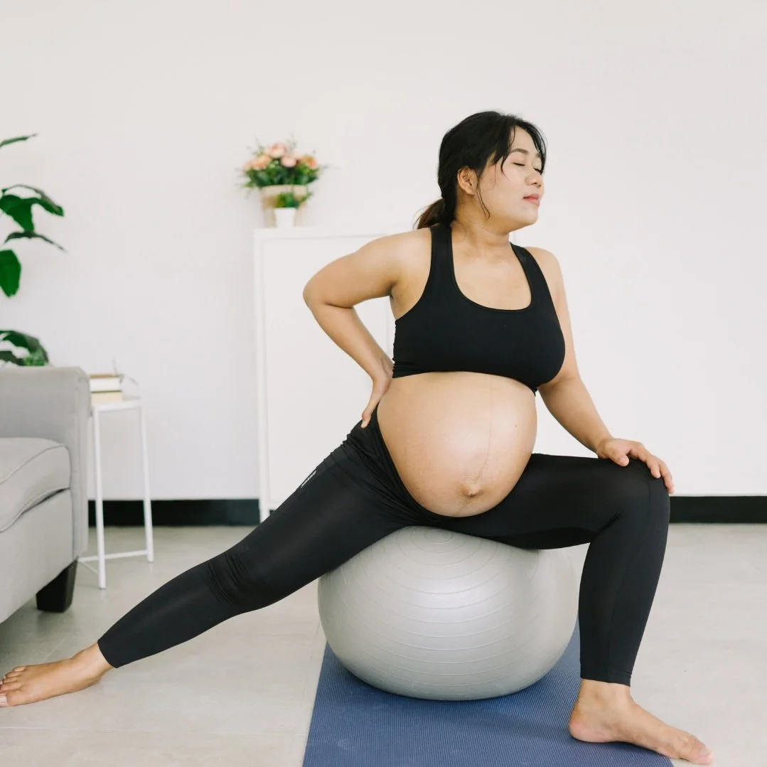 Pregnant woman in black workout attire sitting on an exercise ball practicing prenatal yoga in a living room setting.